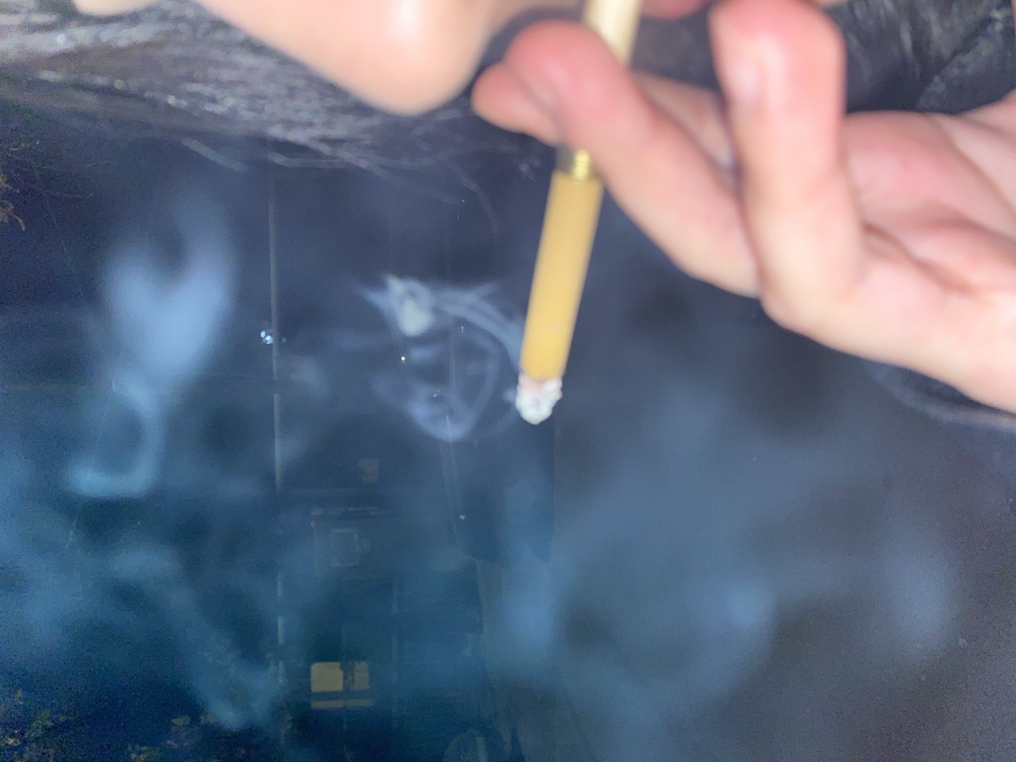 A hand holds a lit cigarette, with smoke billowing out against a blurred, dark background that suggests an outdoor setting. The background includes indistinct building details, possibly indicating an urban environment.