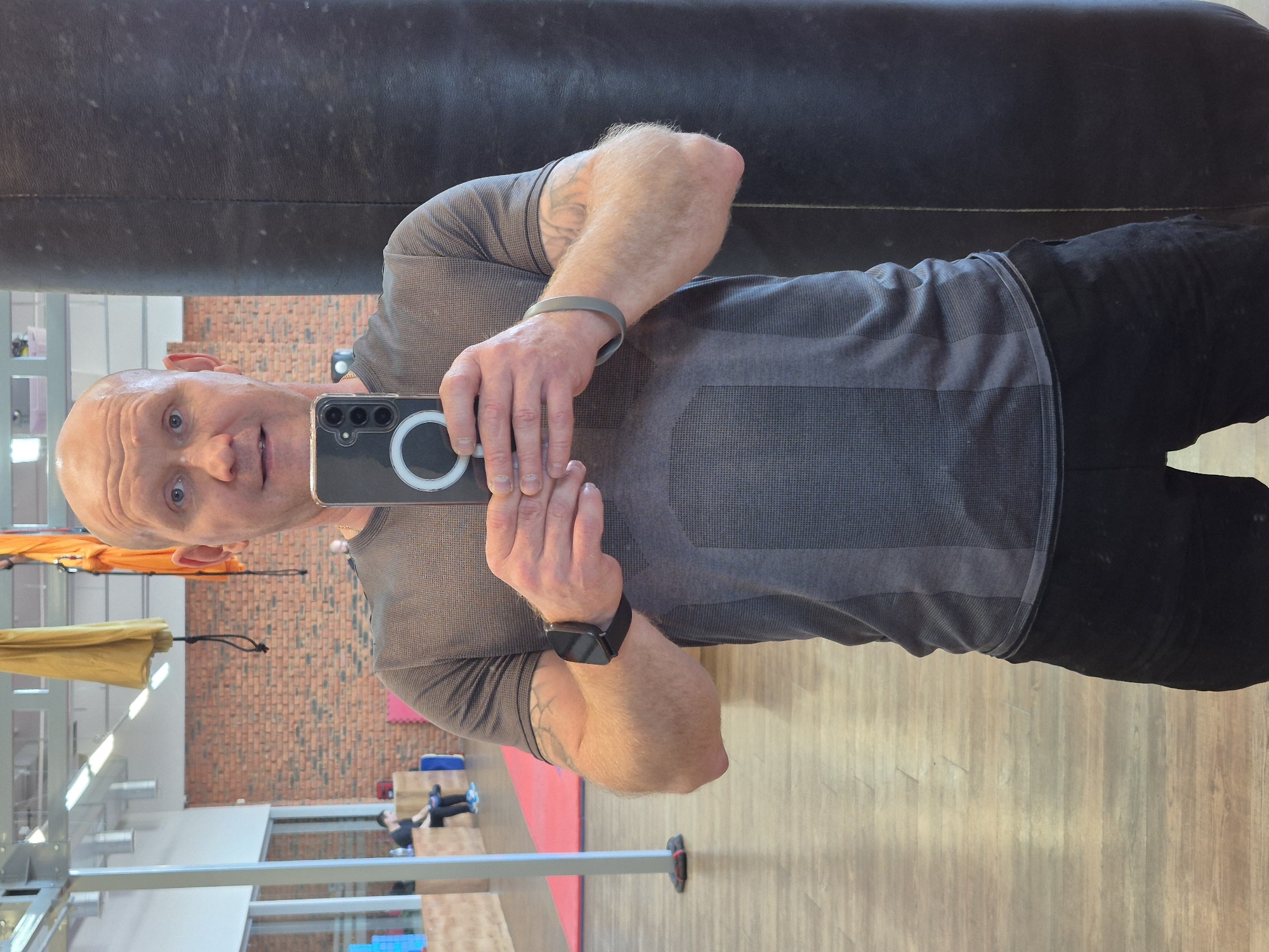 A bald male, mid-thirties to early forties, with light skin and blue eyes, is centered in the image, taking a mirror selfie in an indoor fitness facility in Chaikovsky, Russia. He wears a dark grey short-sleeved athletic shirt with a textured pattern and black shorts. A black smartwatch is present on his left wrist, and a grey silicone wristband is on his right wrist, which also shows an indistinct tattoo. He holds a black smartphone with both hands, positioned in front of his torso, featuring a triple camera array and a white ring-light attachment illuminating its reflection. The man maintains direct eye contact with the camera, exhibiting a neutral to slightly smirking expression.
The background reveals a gym environment with light wood-patterned flooring. A large, dark grey or black punching bag is partially visible on the right side of the frame. In the mid-ground, an exposed red brick wall is visible. The ceiling features metal support structures and rectangular fluorescent light fixtures. Hanging from the ceiling are two fabric hammocks or slings, one yellow and one orange, suggesting aerial fitness equipment. To the left, wooden plyometric boxes or platforms are present, and a red mat is visible on the floor further back. Another male individual, dressed in dark clothing and blue sneakers, is partially visible sitting on a wooden platform in the far left background.