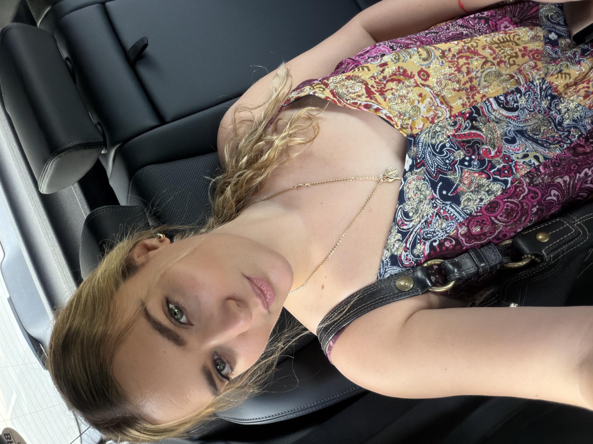 A woman is lying on the black leather back seat of a vehicle. She is wearing a colorful patchwork-style dress with gold detailing and a delicate gold necklace. A black leather purse with a strap rests near her shoulder. The vehicle's interior is clean and well-maintained. The location cannot be determined from the image, and therefore, specifying the city of Miami is not possible.