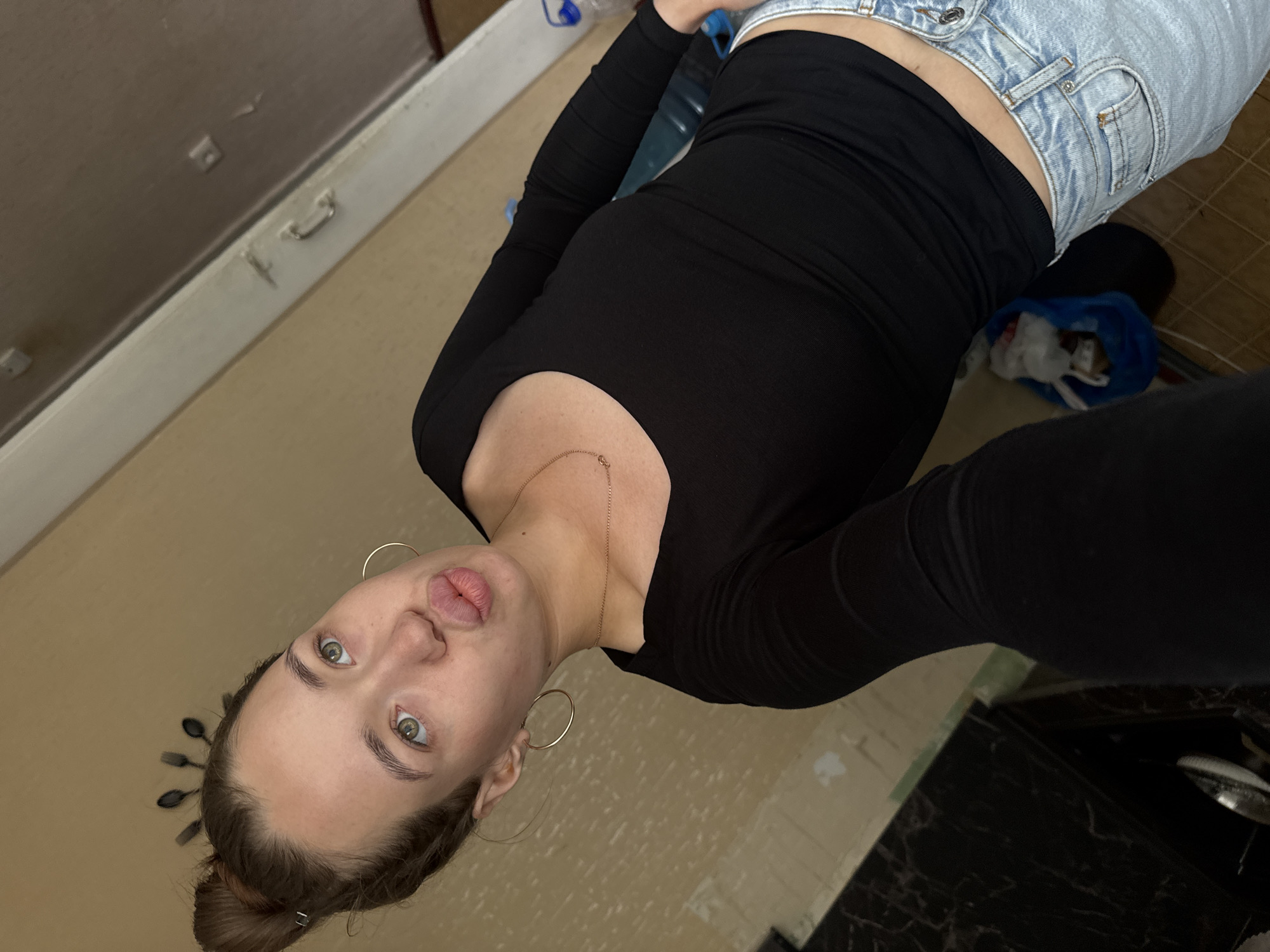 A young woman with her hair in a bun is taking a selfie indoors. She is wearing a black long-sleeved shirt and light-wash jeans. She has large hoop earrings and a thin gold necklace. Her lips are pursed in a duck face. In the background, there is a beige wall and a glimpse of a metallic countertop. The image appears to be taken in a residential setting, possibly an apartment. There is no information to suggest this location is in Chaikovsky, Russia.