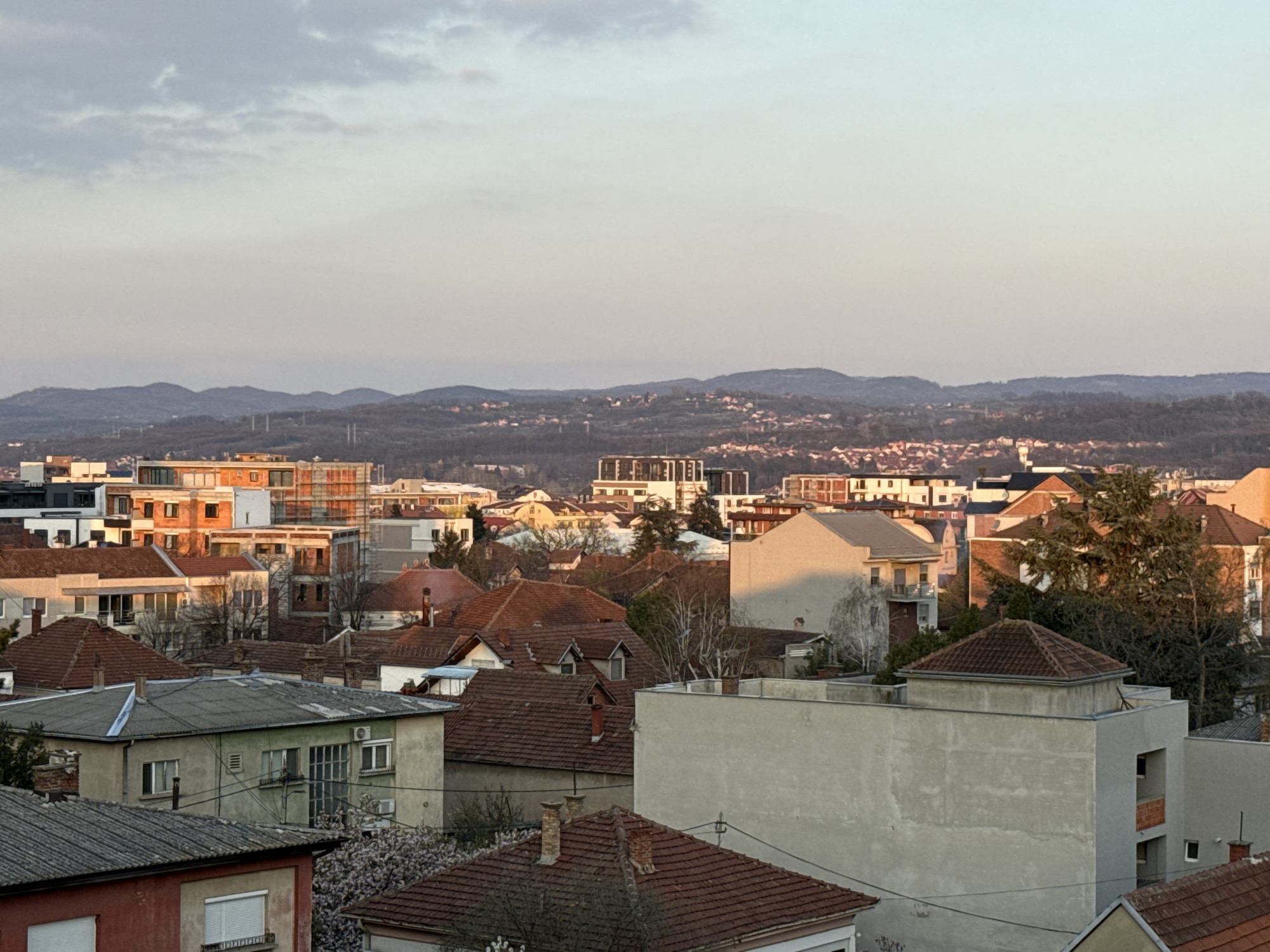 The image presents a cityscape in Kragujevac, Serbia. In the foreground, there are predominantly residential buildings with tiled roofs, with varying architectural styles. Some are older, single-story constructions, while others are taller, multi-story buildings. The buildings are a mix of concrete, brick, and stucco construction.
In the mid-ground, more modern structures are visible. These include apartment buildings with glass balconies and contemporary design features. These structures appear to be of more recent construction.
In the background, the horizon line is formed by rolling hills, likely the surrounding natural landscape of the region. There is a light haze in the distance.
The sky is overcast, with pale, muted colors, suggesting either early morning or late afternoon light. Overall, the scene conveys a mixed urban landscape of old and new architecture, with a natural backdrop. There are no people visible in the image.