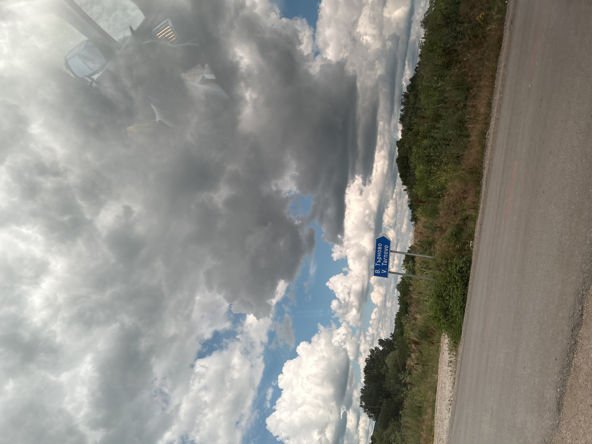 An eye-level, exterior, medium shot shows a cloudy sky above a rural road. A blue directional sign points to the left and reads "V. Tarnovo". The road is gray asphalt with a shoulder of gravel and dry grass. Dense green foliage and trees line the right side of the road. The sky is filled with large, gray and white cumulus clouds, with patches of blue sky visible. Reflections of what appears to be a vehicle's interior are visible in the upper right portion of the image.
