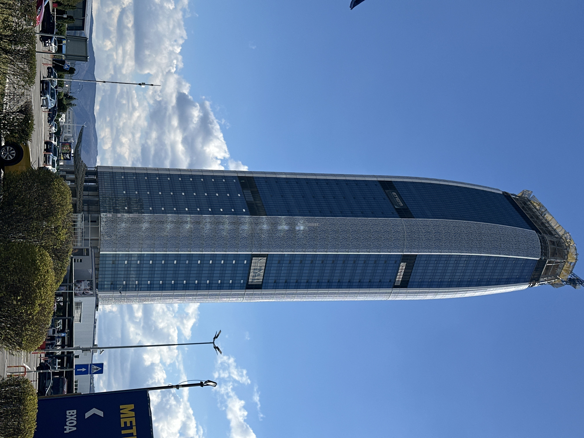 The image shows the Capital Fort building in Sofia, Bulgaria. The building is a modern skyscraper with a curved top and glass facade. It has a grid-like pattern of windows and a section with a perforated metal exterior. The sky is blue with scattered clouds. The foreground shows vegetation, streetlights, cars, and a sign with the word "METRO" in Cyrillic script.