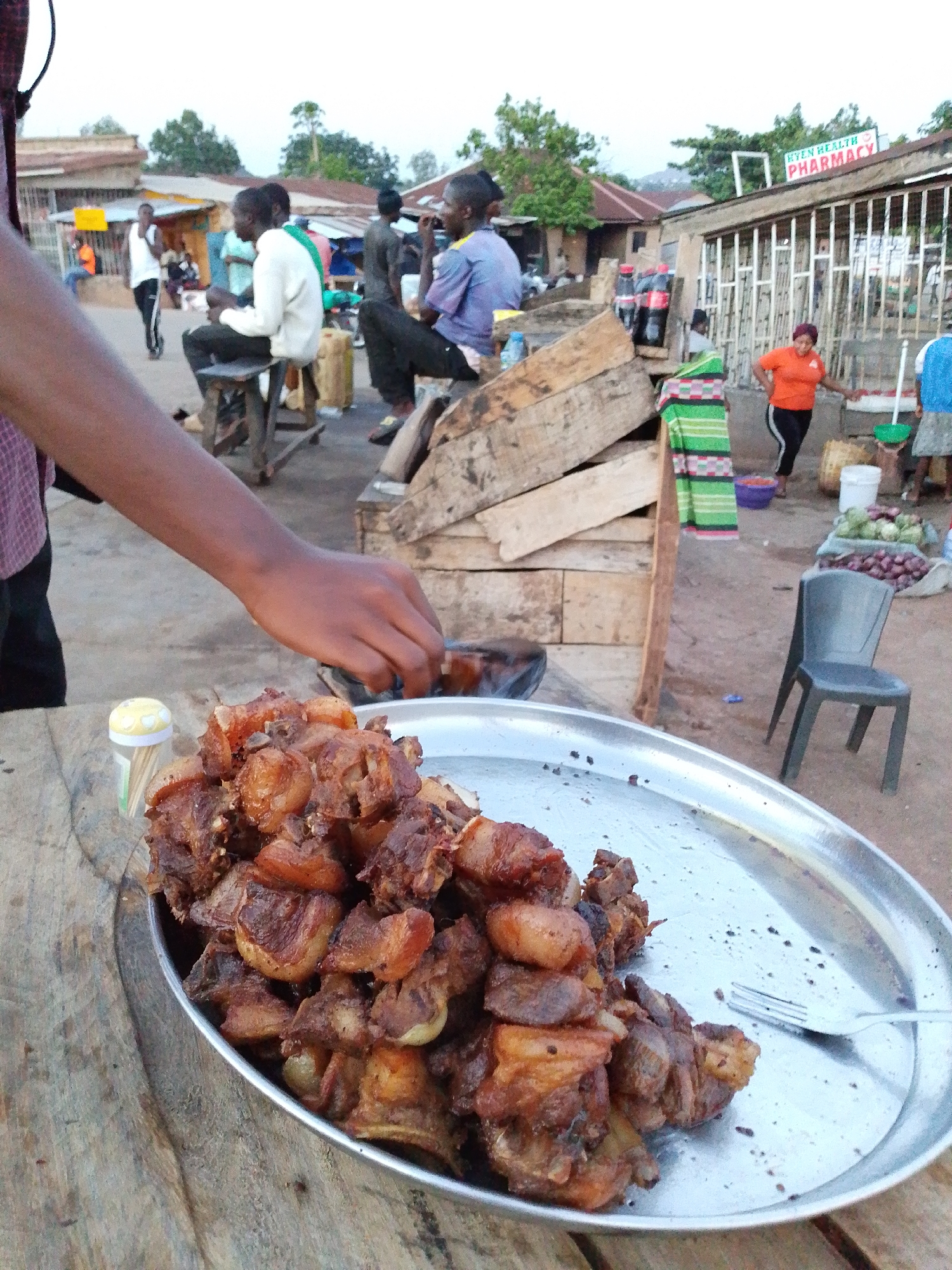 The image depicts a scene in what appears to be a marketplace in Kuru Dungus, Nigeria. In the foreground, a large silver platter filled with chunks of fried meat sits on a rough wooden table. A hand reaches towards a small bowl or container next to the platter. A container of toothpicks is also visible.
In the background, people are seen sitting and standing around wooden structures. One structure appears to be a makeshift seating area. A woman in an orange shirt stands near a stall selling produce, including onions and cabbage. A sign reads "HYEN HEALTH PHARMACY." Other people are milling about in the marketplace. The environment appears to be a somewhat dusty outdoor area.