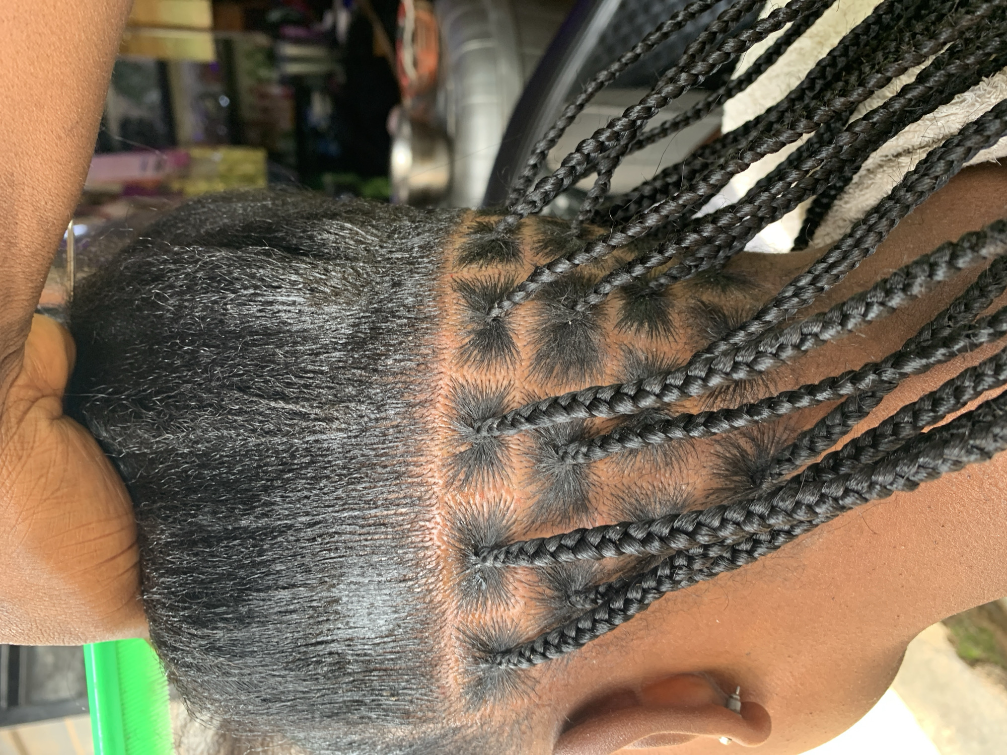 The image depicts a close-up, overhead view of the back of a person's head undergoing hair braiding in Rafin Par, Nigeria. The subject, viewed from behind and the left profile, has dark skin and coarse, black hair. Numerous small box braids are partially completed, emerging from precisely defined square partings on the scalp. Several finished braids extend downwards, while unbraided sections of hair are held by a dark-skinned hand and forearm at the top of the frame, indicating the presence of a braider. A portion of a green comb is visible on the upper left. The subject's left earlobe features a small, silver stud earring. A light-colored towel is draped around the neck area. The background is softly blurred, revealing indistinct interior elements such as shelving, metallic bowls, and furniture, suggesting a salon or domestic environment.