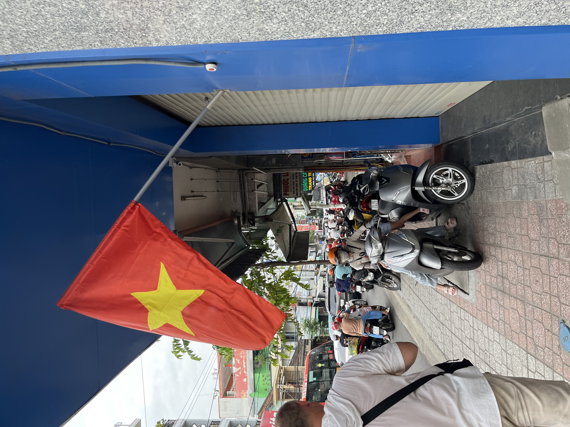 The image depicts a street scene in Nha Trang, Vietnam, with a prominent Vietnamese flag, red with a yellow star, extending from an overhead fixture. The flag is partially obscuring a blue building facade. The sidewalk is paved with interlocking bricks in a geometric pattern. In the background, a busy street is visible with numerous motorcycles, cars, and a red bus, indicating moderate traffic. Several individuals are seen on motorcycles, some wearing helmets. The environment suggests a commercial area with shops lining the street. The overall scene captures a slice of everyday life in a Vietnamese city.