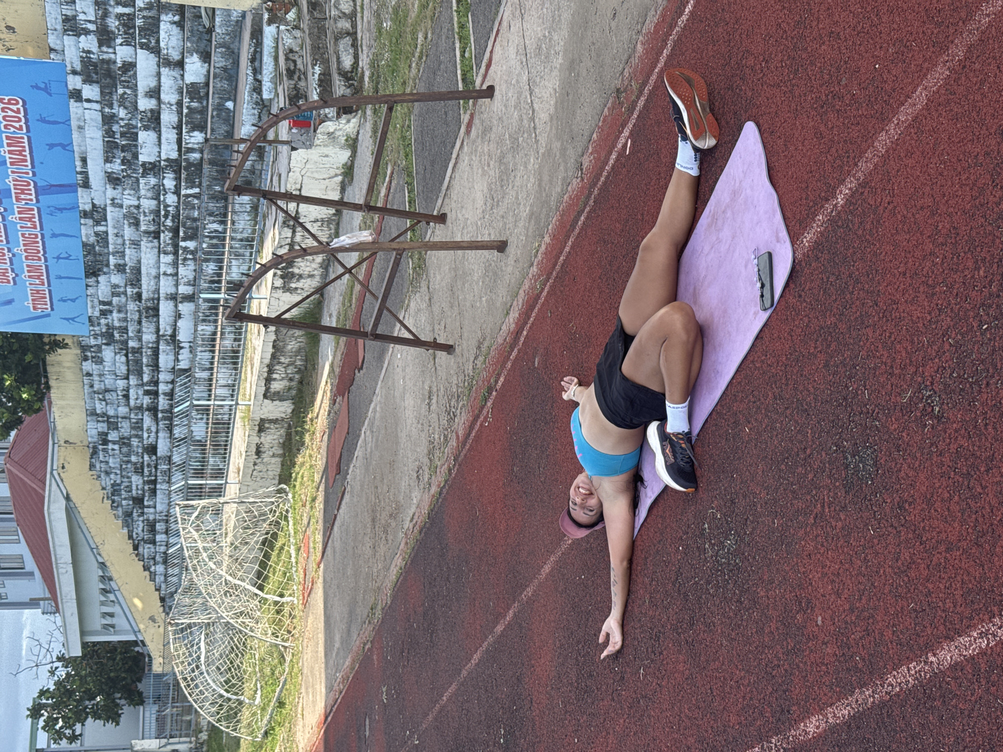 A woman lies on a purple yoga mat on a red running track, with her arms and legs spread wide. She is wearing a blue sports bra, black shorts, a pink baseball cap, and athletic shoes. She is smiling at the camera. In the background, there are stadium bleachers, a soccer goal, and a building. The setting appears to be a stadium in Phan Thiết, Vietnam, possibly during the day.