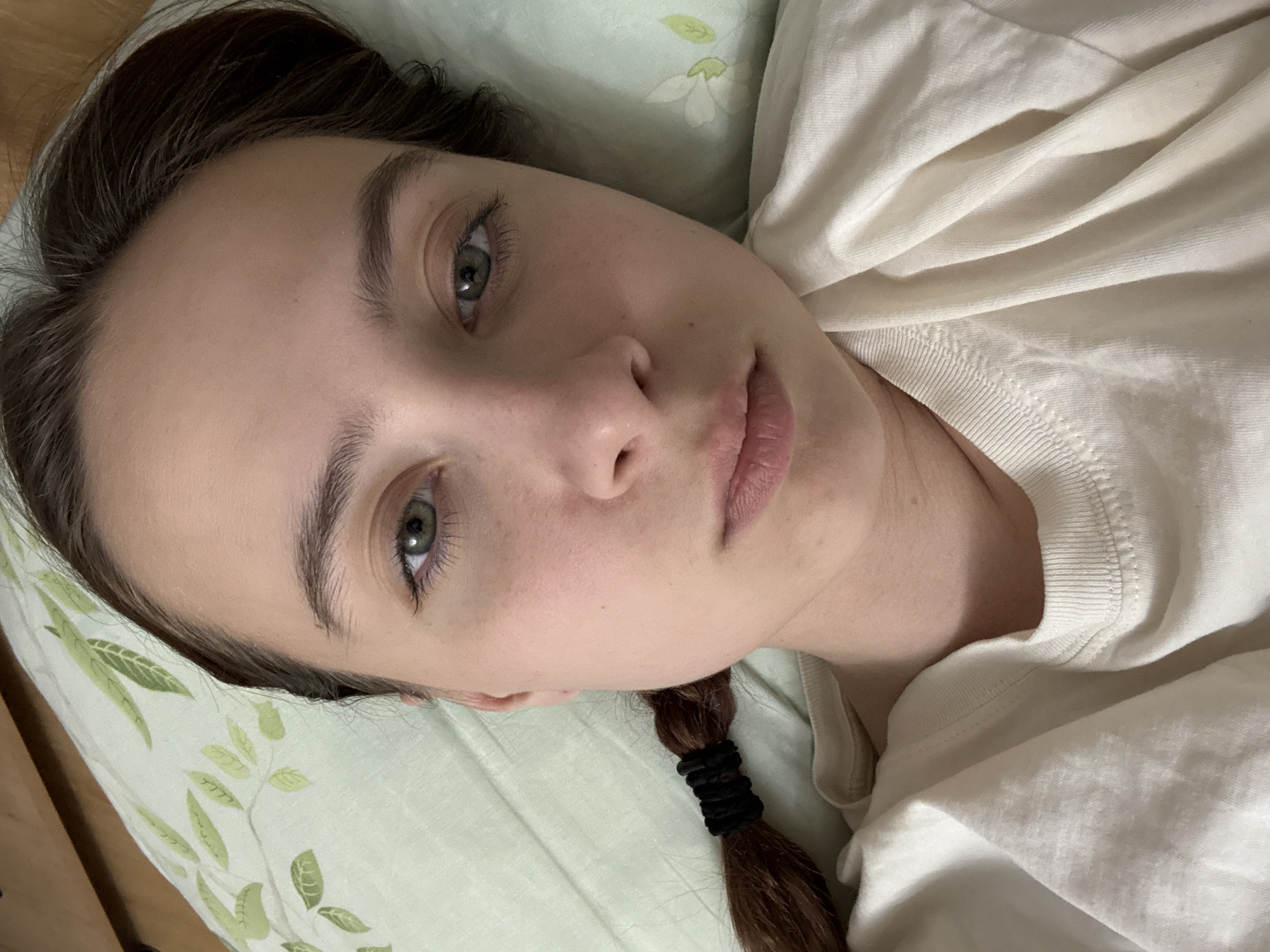 A young woman with brown hair is lying on a pillow covered with a light green floral pattern. She is wearing a white shirt. The woman's hair is partly braided and secured with a black hair tie. Her eyes are green. The image provides no evidence that allows the determination of the location as Berezniki, Russia.