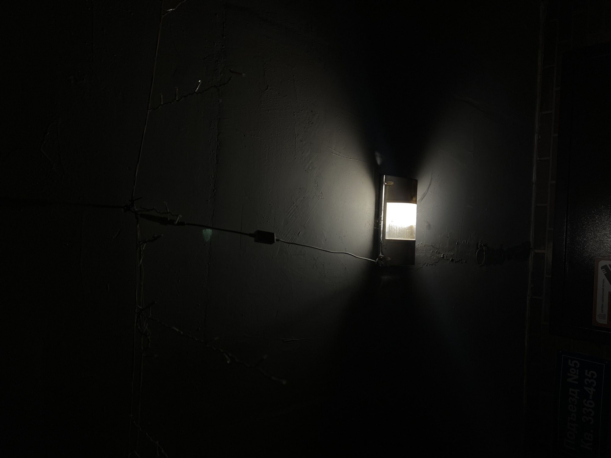 A dim light illuminates a textured black wall in what appears to be the entranceway of an apartment building in Lyubertsy, Russia. A light fixture is recessed into the wall with a metal frame. A wire runs horizontally along the wall to the left of the light fixture, possibly connected to it. To the right of the light, a darker area suggests a doorway or entrance. A small sign in Cyrillic is visible in the lower right corner, partially obscured by darkness, and likely indicates the apartment building entrance number. The wall is uneven and cracked in places. There are no people visible in the frame.