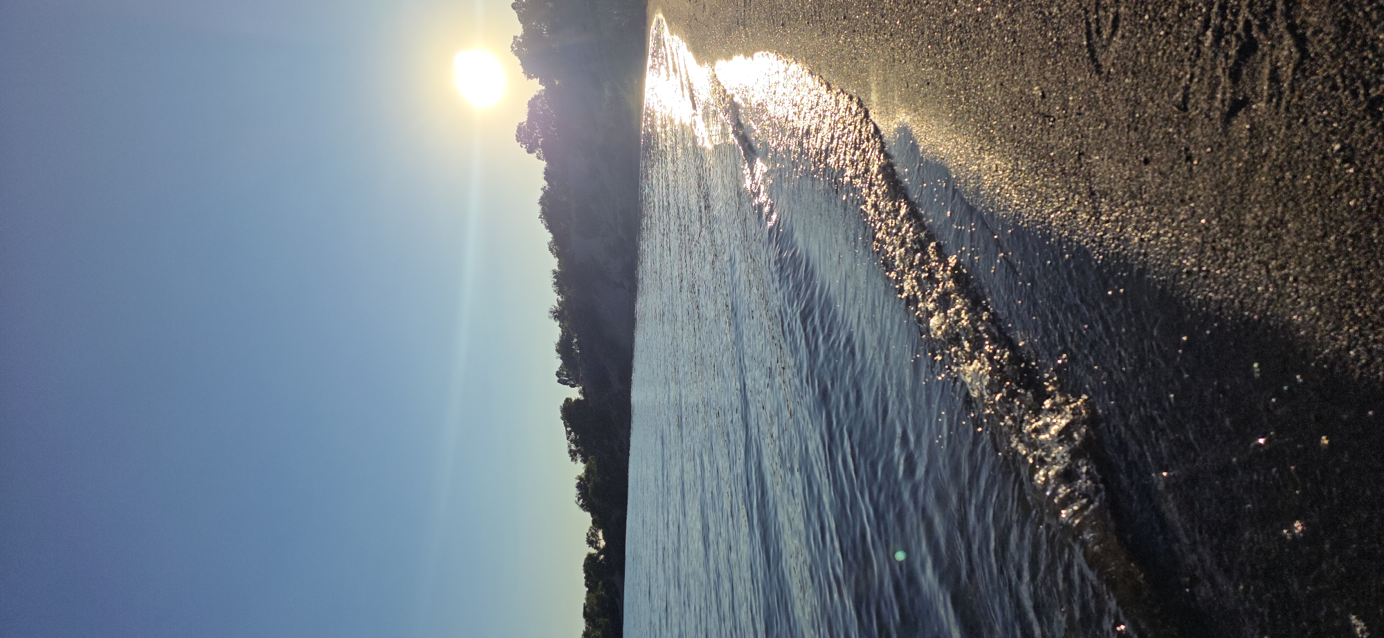 This is a photograph of a beach at sunset on Issyk-Kul Lake in Kyrgyzstan. The sun is low on the horizon, casting a golden glow on the water and sand. The waves are gentle, lapping at the shore. Trees and vegetation line the distant bank. The sky is a clear blue, transitioning to a lighter hue near the horizon. The image captures a serene and natural environment.