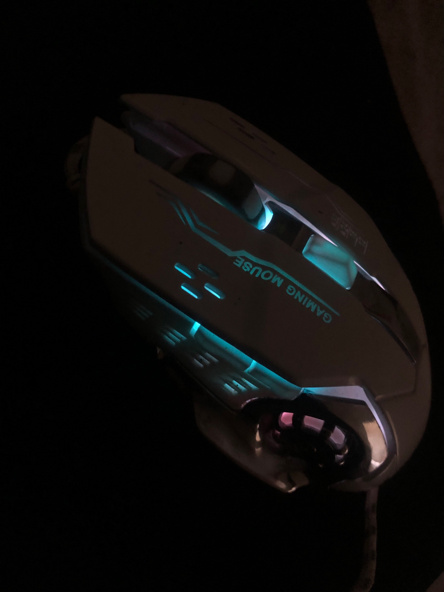 The image shows a white gaming mouse with blue and pink LED lights on a dark background. The mouse has the words "GAMING MOUSE" printed on its surface in a stylized font. The mouse is positioned at an angle, with its cord visible at the bottom. The image does not depict any identifiable location in Saidan, Pakistan, nor does it contain any people or indicate any specific actions or interactions.