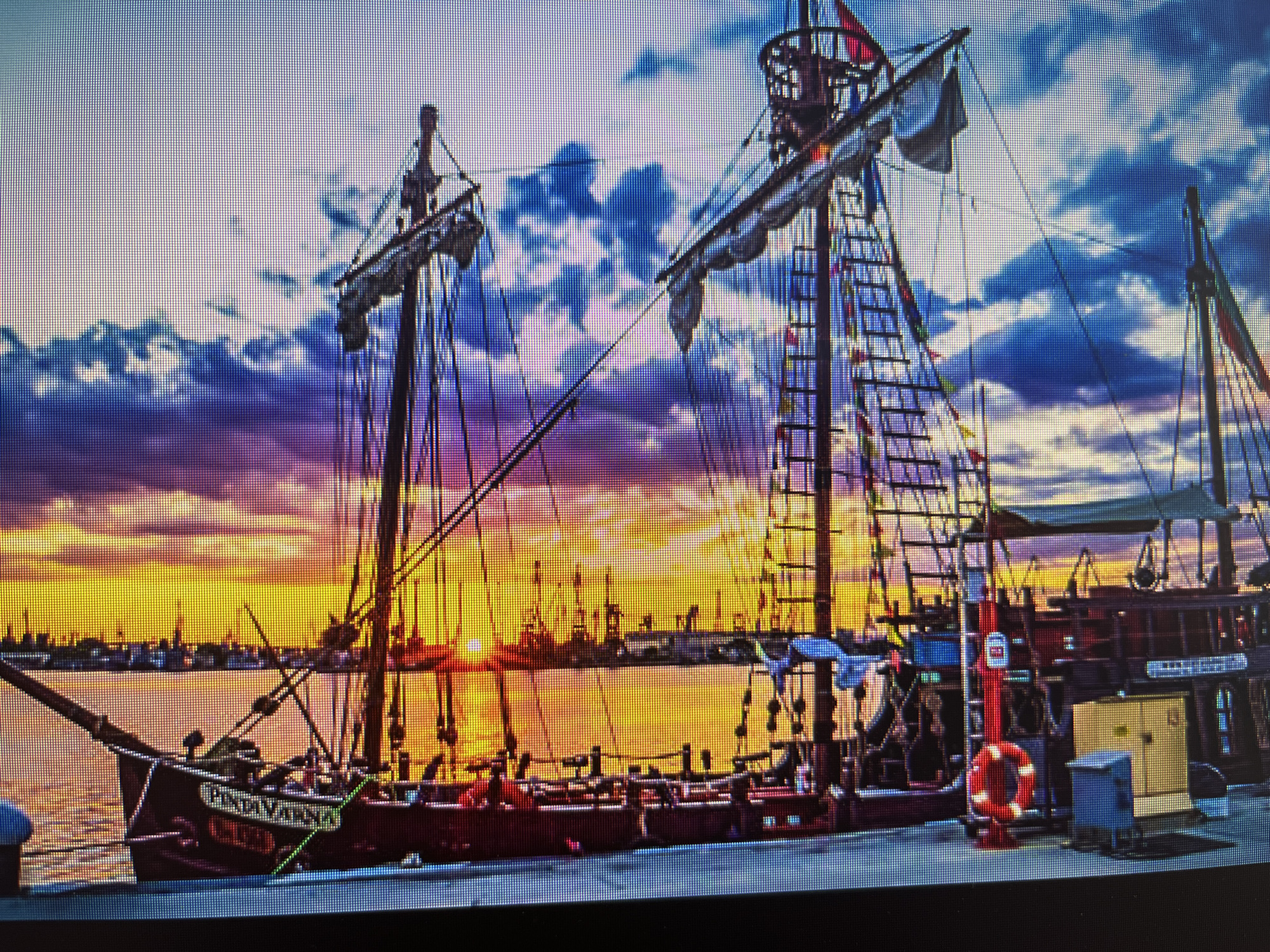 The image depicts a replica of the Pinta ship, docked in Varna, Bulgaria, at sunset. The sky is a mix of purple, blue, and orange hues. The ship's masts and rigging are silhouetted against the colorful sky. In the background, a row of city buildings and port cranes are visible along the waterfront. Closer to the ship, a yellow utility box and a red life preserver are observed on the dock. There are no visible people. The ship bears the inscription "PINTA VARNA."