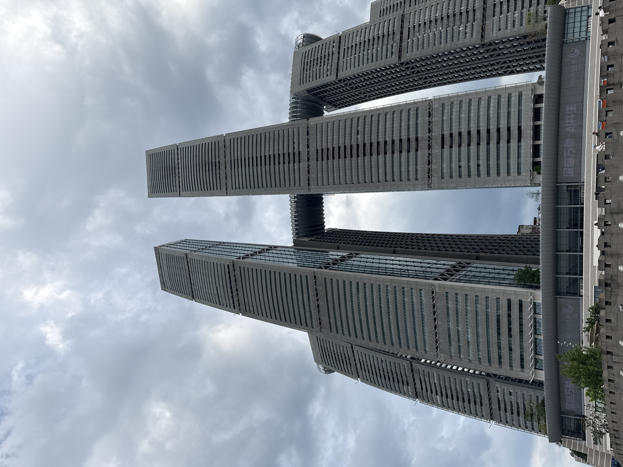 The image shows a low-angle, wide shot of the Raffles City Chongqing, a complex of skyscrapers in Yuzhong District, China. The buildings are tall and slender, with a dark gray facade and horizontal window bands. Two of the tallest buildings are connected by a horizontal skybridge that resembles a ship or a crystal. The skybridge is enclosed and appears to have a glass facade, offering panoramic views. The buildings are set against a cloudy sky, with the clouds being a mix of white and gray, suggesting an overcast day. At the base of the buildings, a lower-level structure with a modern design is visible, featuring large illuminated panels with Chinese text and logos. In the foreground, there is a grey stone wall with decorative elements, and several individuals are visible standing on or near the wall, appearing to observe the architecture. Some greenery, including trees, is also present at the base of the skyscrapers.
