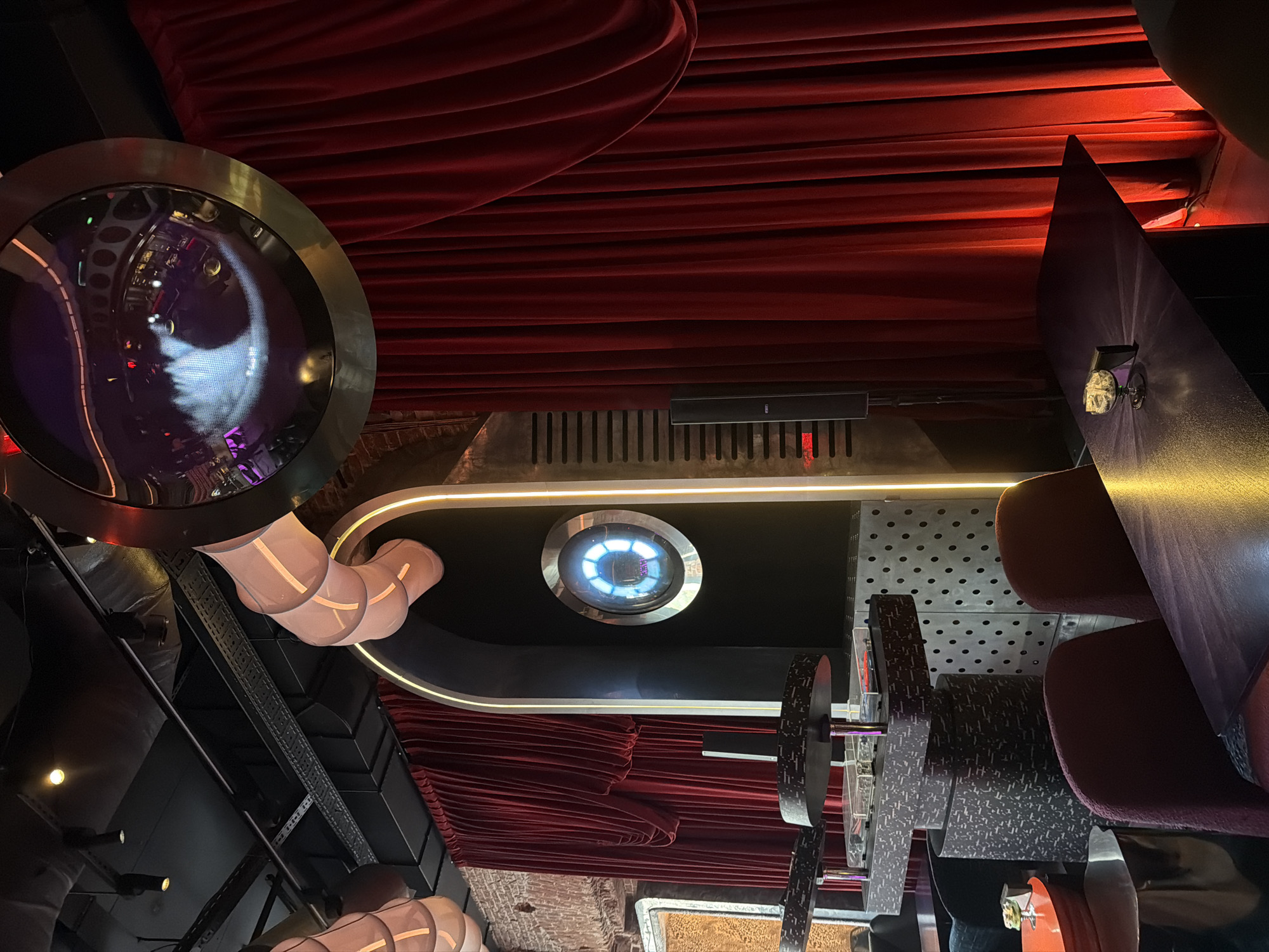 The image depicts the interior of a venue in Moscow, Russia, characterized by a theatrical, steampunk-inspired aesthetic. Red velvet drapes cascade from the ceiling, contrasting with the dark, industrial elements. A large, circular metallic structure resembling a porthole dominates the left side of the frame, reflecting interior lights. A central, circular portal with illuminated concentric rings is set into a black panel with a curved, illuminated border. Below this, more red drapes are visible. To the right, an arrangement of decoratively textured panels, wheel-shaped objects, and partially visible furniture, including chairs upholstered in maroon, suggests a seating area. The overall impression is that of a themed entertainment or dining establishment with a focus on dramatic design.
