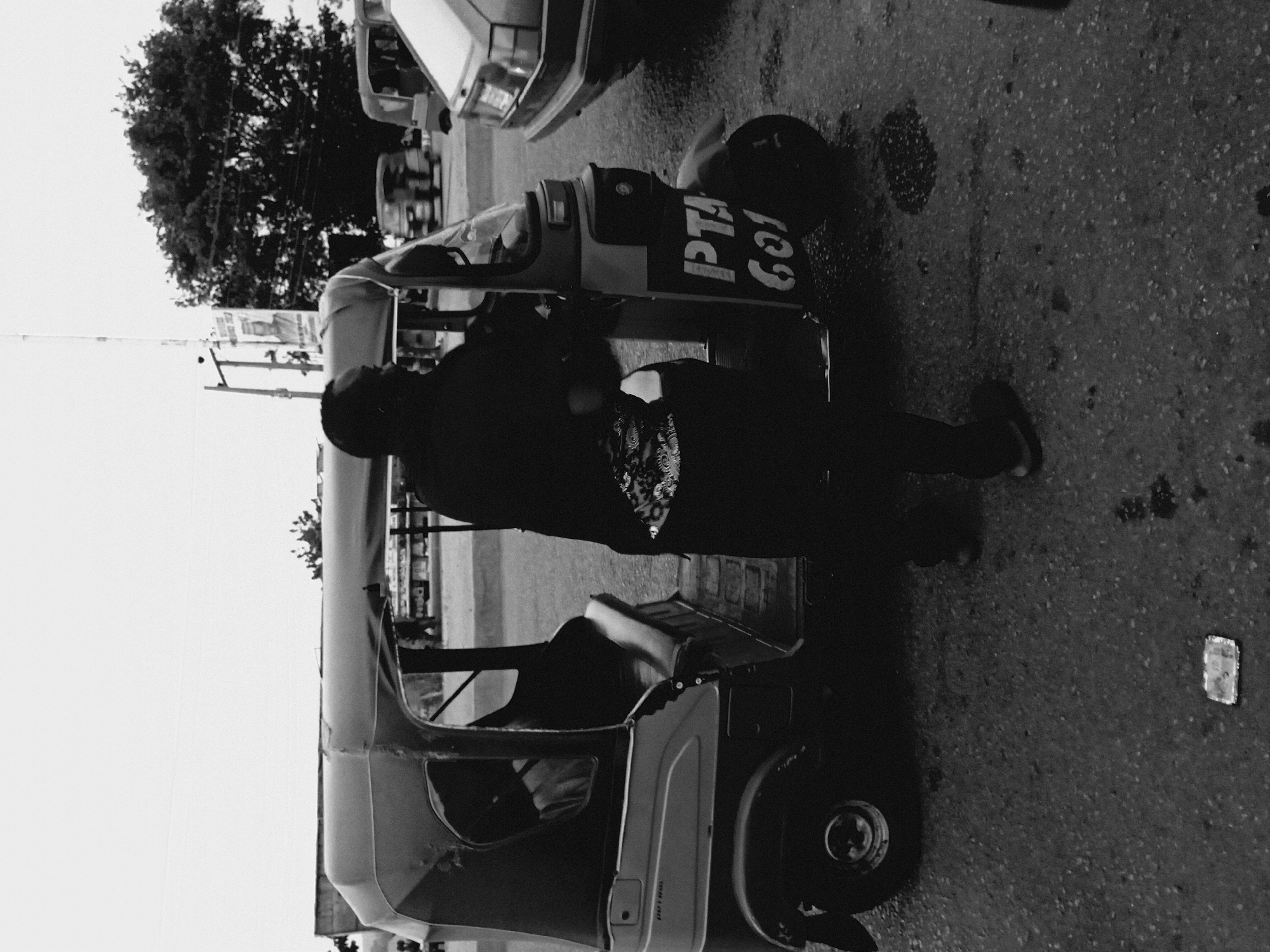 A black and white photograph depicts a person standing next to a parked auto rickshaw on a street in Bukuru, Nigeria. The person is facing the auto rickshaw and appears to be interacting with it. The auto rickshaw is parked on the side of the road, and there are other vehicles and buildings visible in the background. The road appears to be paved and wet.