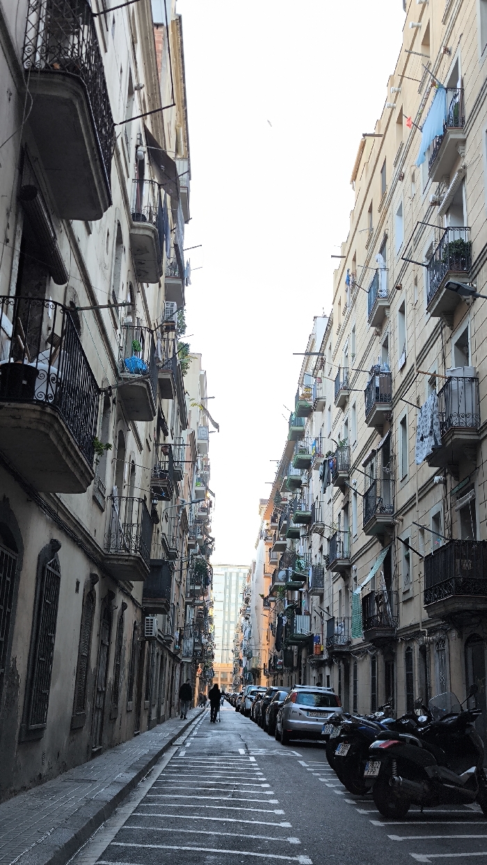The image captures a perspective view down a narrow, bustling street in what appears to be a European city. Tall, multi-story residential buildings line both sides of the street, creating a deep urban canyon that recedes into the distance. The architecture features numerous balconies with wrought-iron railings, some adorned with potted plants, and many displaying clothes hanging out to dry, indicating an active residential environment. Air conditioning units are also visible on several building facades.
The buildings on the left side of the street appear to be an older, muted beige or greyish tone, with arched window details and heavy cornices. The buildings on the right side are a lighter, warmer cream or light yellow color, also with numerous balconies. The sky above is a bright, almost featureless white, suggesting strong daylight, though the street level itself is somewhat shadowed, especially in the foreground.
The street is paved, with a sidewalk running along the left. The road features distinctive white horizontal markings that segment the street, possibly indicating parking zones or speed calming measures. Along the right side of the street, a line of cars is parked bumper-to-bumper, followed by several motorcycles and scooters parked closer to the foreground. License plates are visible on some vehicles; a silver car has "2468 GSV" visible, and a scooter in the foreground displays "7722 FPX".
Two figures can be seen walking in the distance along the left side of the street; one appears to be pushing a stroller or cart. The overall scene conveys a sense of everyday urban life in a densely populated area.