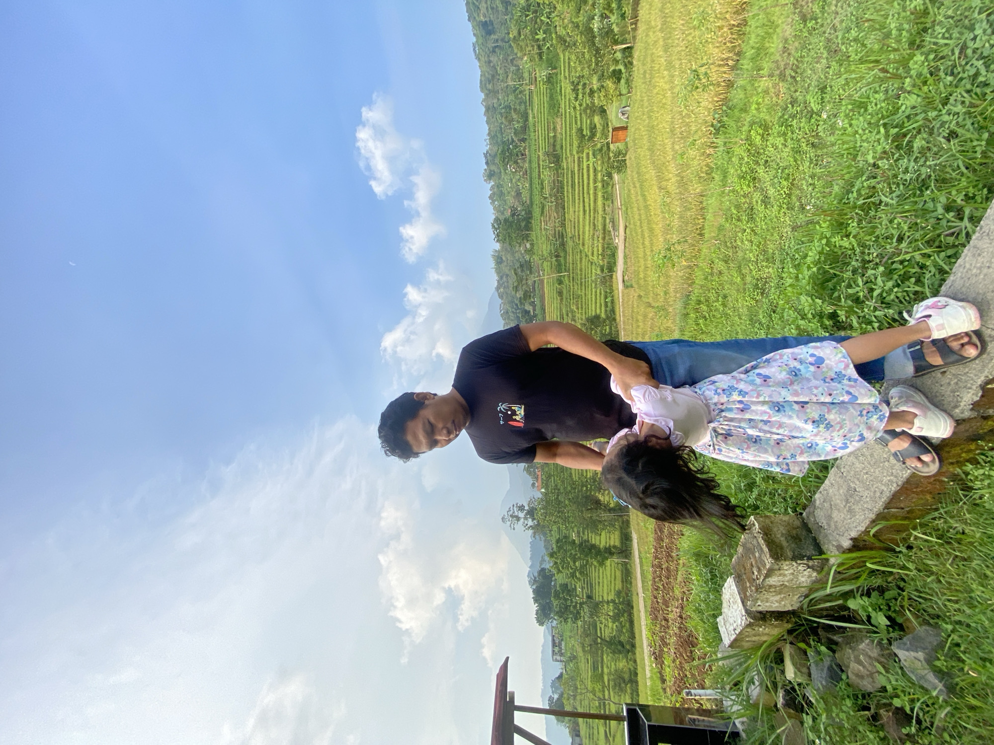 In Penanggungan, Indonesia, a man in a black shirt and blue jeans is holding a young girl in a floral dress. She is leaning back, with her head and hair dangling over the edge of a concrete barrier. The background includes rice paddies, green foliage, a hillside, and a blue sky with scattered clouds.