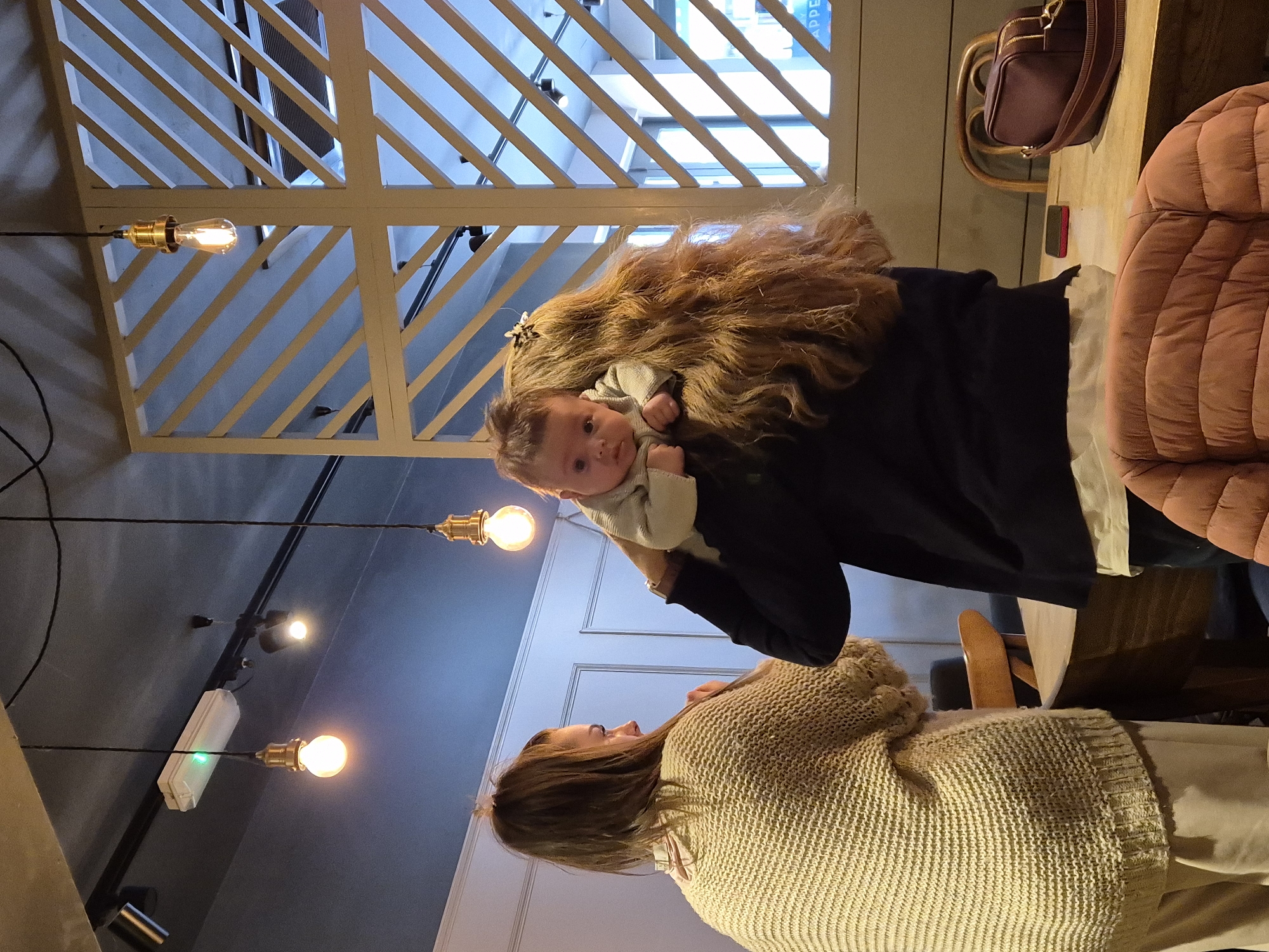 Here is a detailed description of the image:
The image is taken indoors, likely in a cafe or restaurant. A woman in a beige knitted sweater stands facing another woman who is holding a baby up in the air. The woman holding the baby is wearing a black jacket and has long hair. The baby is looking at the camera. The scene is lit by multiple exposed bulb light fixtures hanging from the ceiling. The ceiling has a decorative lattice design. A brown handbag rests on a table next to a pink quilted seat. A wooden chair is also visible. The image appears to have been taken in West Suffolk, United Kingdom.