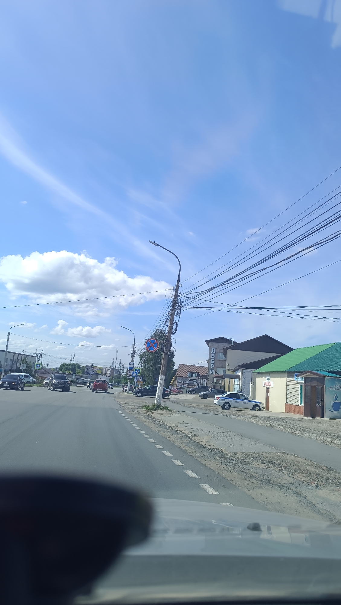 The image captures a view from a vehicle traveling down a street in Chaikovsky, Russia, on a clear day with scattered clouds. The road has distinct white dashed lines marking the lanes and a gravel shoulder. On the right side of the road, there are several buildings, including one with a green roof and signs that appear to be for a pharmacy and a cafe. Further down the street, a police car is parked. A prominent utility pole with a multitude of wires stands near the intersection, leaning at an angle and adorned with traffic signs. On the left side of the road, other vehicles are visible, including cars and what appears to be an SUV. Buildings and more utility poles can be seen in the background.