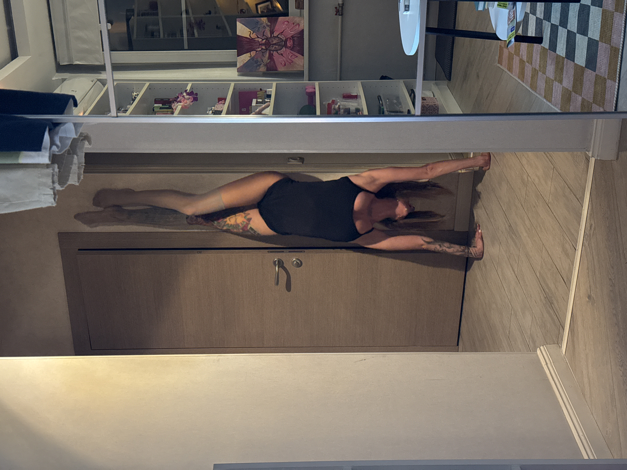 The image captures an indoor scene, likely an apartment, with a woman performing a handstand against a wall. She is wearing a black leotard and has tattoos on her arms and legs. The environment includes a wooden door, a floor with a wood-grain pattern, and a large mirror reflecting part of the room. Beyond the mirror, white bookshelves filled with various items, a small white table, and a rug with a checkered pattern are visible. The lighting suggests it is daytime. The scene is devoid of any explicit indicators of its location within Moscow, Russia.