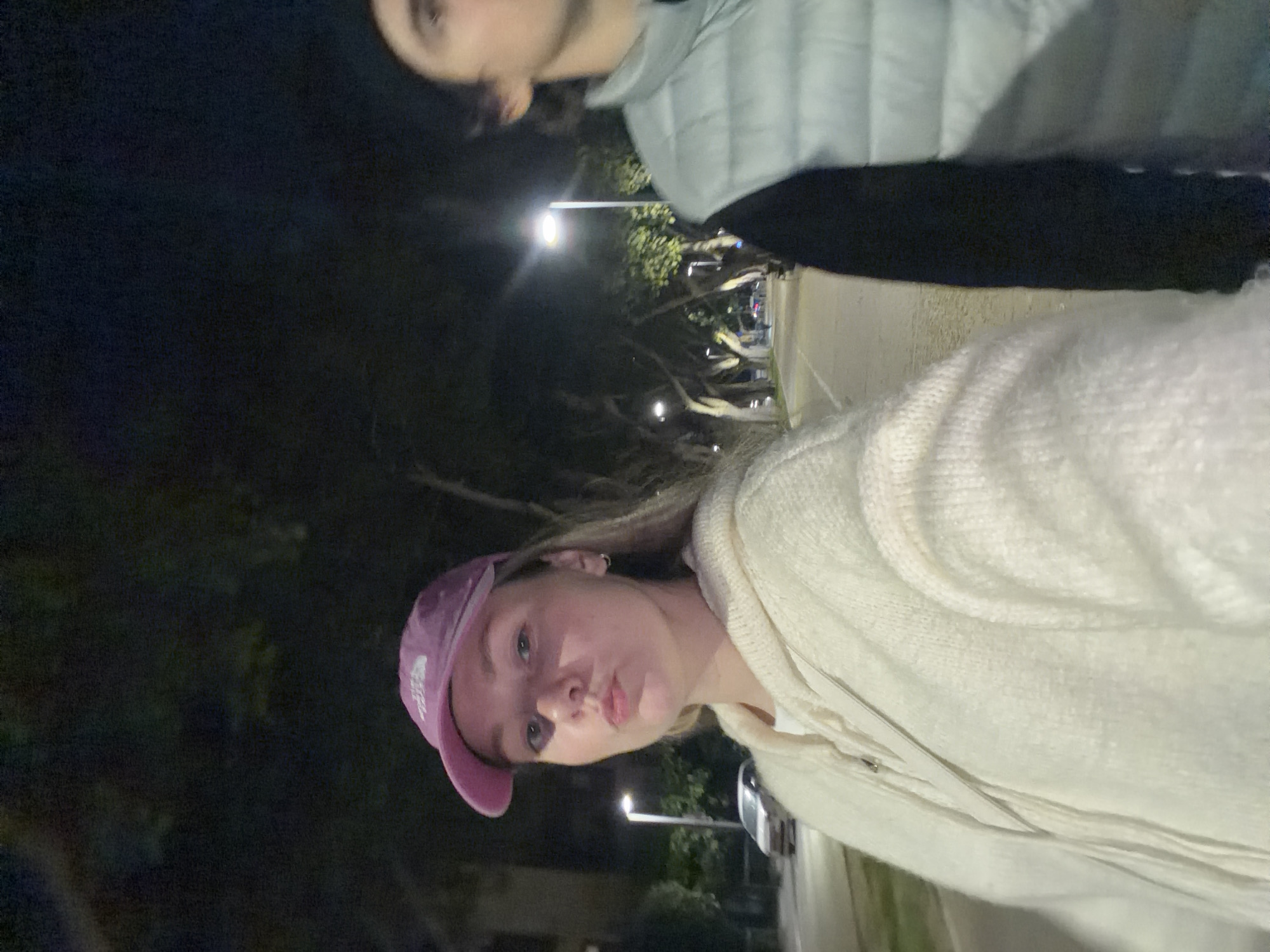 The image depicts two individuals, one wearing a pink cap and a white hooded sweatshirt, posing for a selfie, while the other, dressed in a grey puffer jacket and black pants, stands behind them. The setting appears to be outdoors at night, with street lights illuminating a roadway lined with trees. The environment suggests an urban area. Given that the location of the city is Tel-Aviv, Israel, it's likely the image was taken within this city's boundaries.