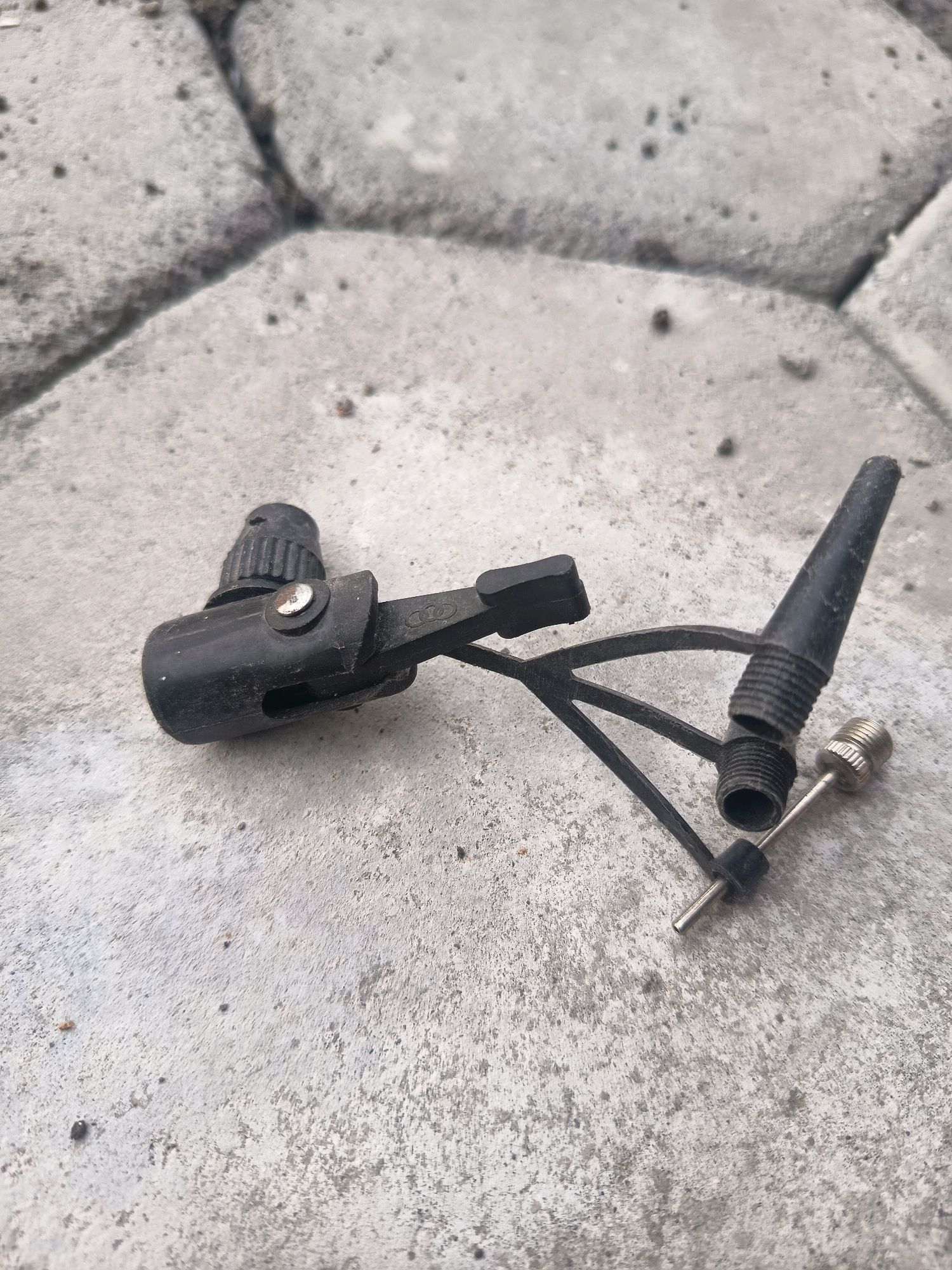 A black plastic bicycle pump adapter is situated on a textured concrete surface. The adapter has a ribbed nozzle at one end, a lever mechanism, and a small metal needle attached by a thin black wire. The concrete surface appears to be part of an outdoor pavement, with visible seams between rectangular slabs. The lighting suggests it is daytime, with shadows casting slightly. The specific location of Sidorejo, Indonesia, is not visually discernible from the image itself. There are no people or actions depicted.