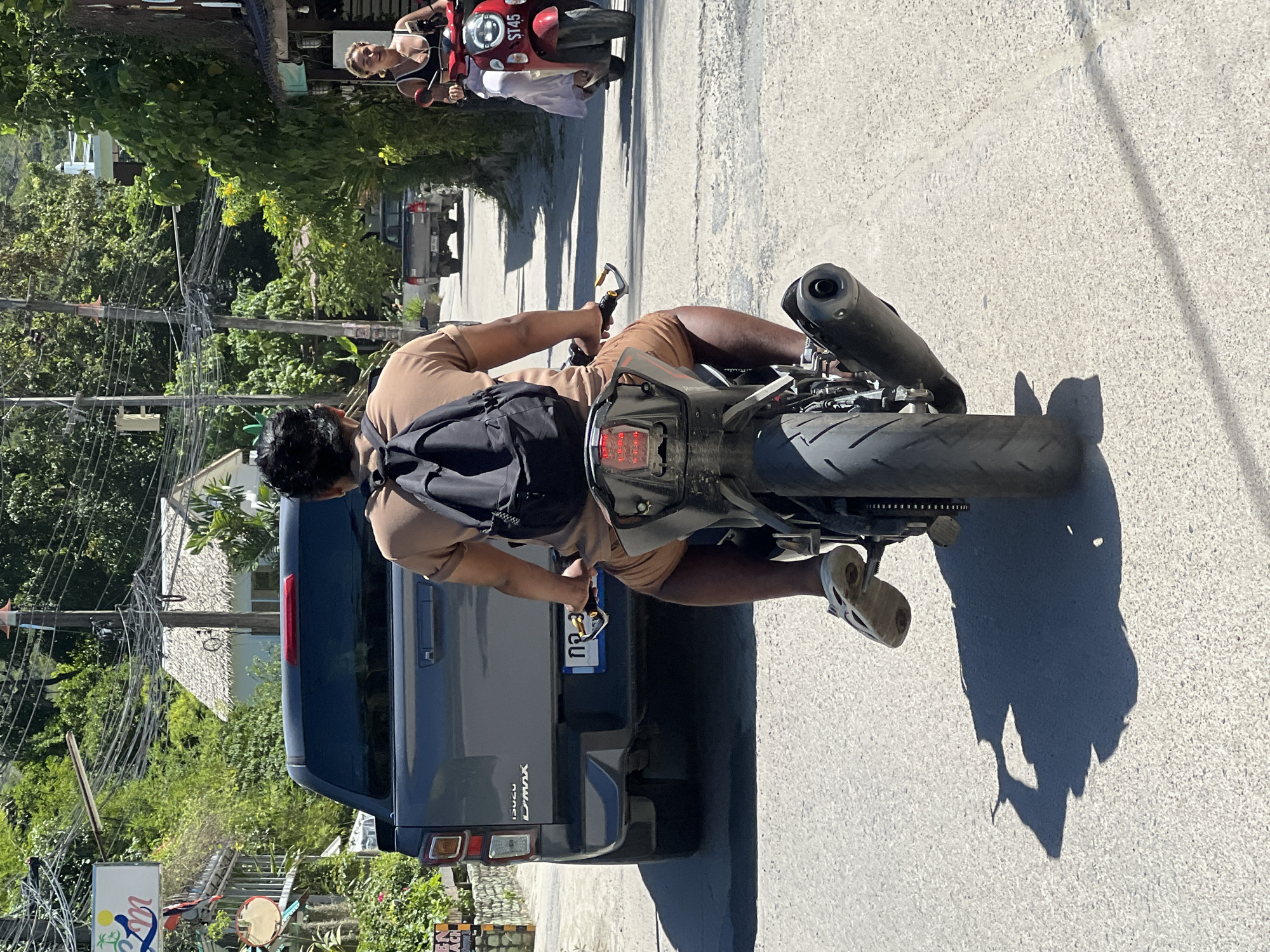 The image depicts a sunny day in a street scene, likely in ตำบลเกาะพะงัน, Thailand, characterized by lush greenery, buildings with thatched roofs, and power lines overhead. In the foreground, a person rides a motorcycle from behind. They are wearing a tan t-shirt, brown shorts, and light-colored sandals, with a black backpack on. The motorcycle is black and has a prominent exhaust pipe. Ahead of the motorcycle, a dark gray Isuzu D-Max pickup truck is visible, with a license plate that appears to have Thai script. To the right of the pickup truck, further down the road, a woman with blonde hair is riding a red scooter, wearing a black tank top and a light-colored skirt, and appears to be looking towards the viewer. The road surface is asphalt, and shadows are cast across it, indicating a bright sun. Various signs and businesses are present along the sides of the street, contributing to a vibrant, tropical atmosphere.