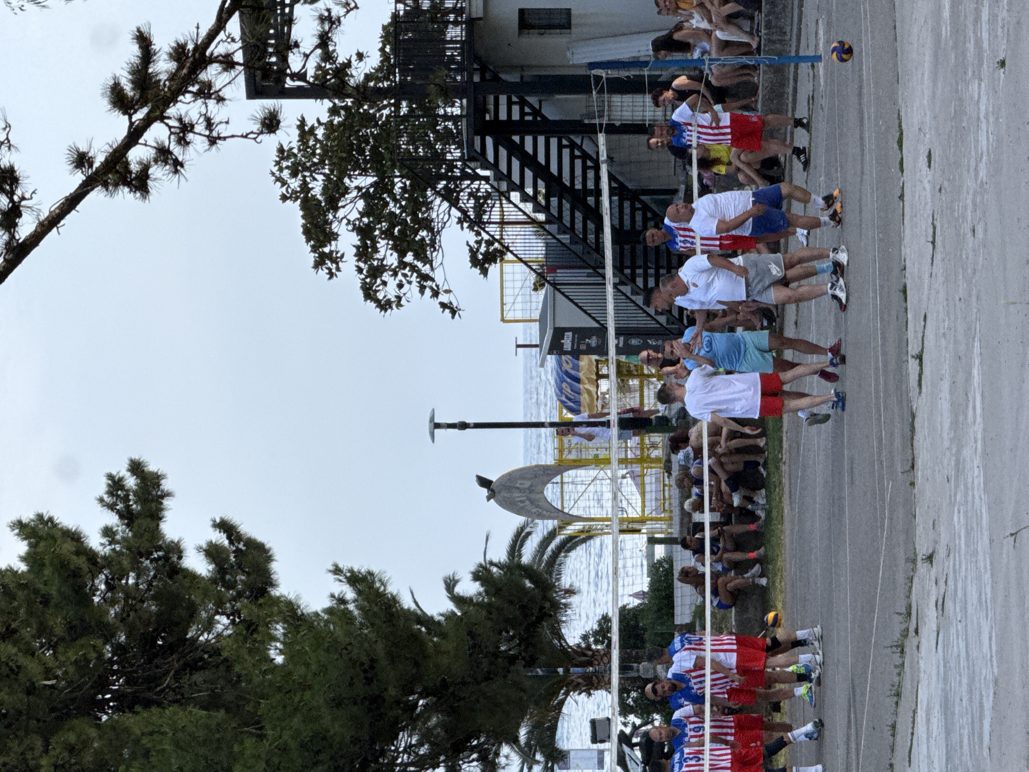 In Budva, Montenegro, a group of men are playing volleyball on an outdoor court near the sea. The court appears to be made of asphalt, with a volleyball net stretched across the middle. Several players are actively engaged in the game, some in motion while others stand ready. Spectators are seated or standing on the sidelines, some under an umbrella for shade. In the background, the Adriatic Sea is visible, with a palm tree and other vegetation framing the scene. A building with an external staircase is also present, suggesting a beachfront facility. The overall atmosphere is one of casual recreation and community engagement on a warm day.