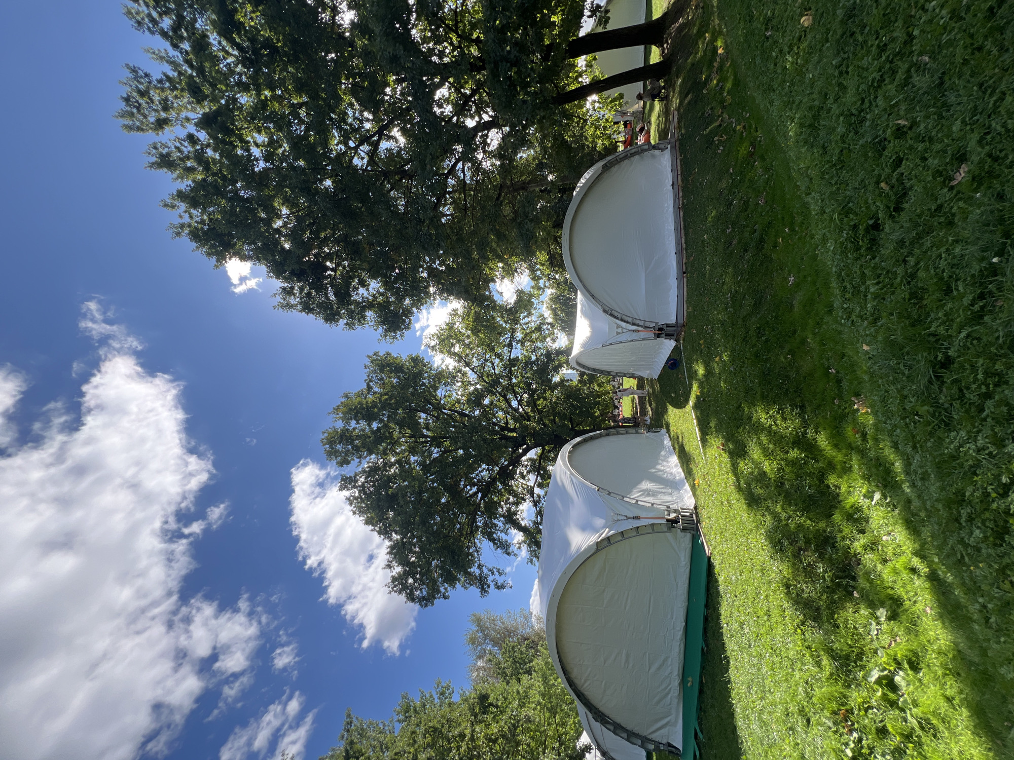 The image depicts a sunny outdoor scene with several white, arched tents set up on a grassy area. The tents appear to be large and modular, with metal framing visible at the entrance. They are positioned amongst mature trees with lush green foliage. The sky above is a vibrant blue with scattered white clouds, suggesting fair weather. The ground is covered in green grass, with some fallen leaves scattered about, indicating it might be autumn. There are a few people visible in the background, including one person sitting on a chair near a tent. The overall impression is a temporary setup in a park-like setting, possibly for an event or outdoor accommodation. No specific landmarks or architectural features that would definitively place this scene in Moscow, Russia, are present in the image.