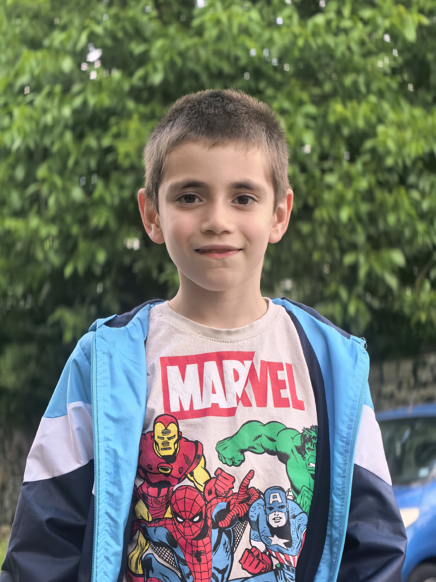 A young boy stands outdoors. He wears a Marvel t-shirt featuring Iron Man, Hulk, Spider-Man, and Captain America, along with a blue, white, and navy jacket. Behind him is a blurred backdrop of green foliage. A portion of a blue vehicle is visible to the right. The location is Unknown, Unknown.