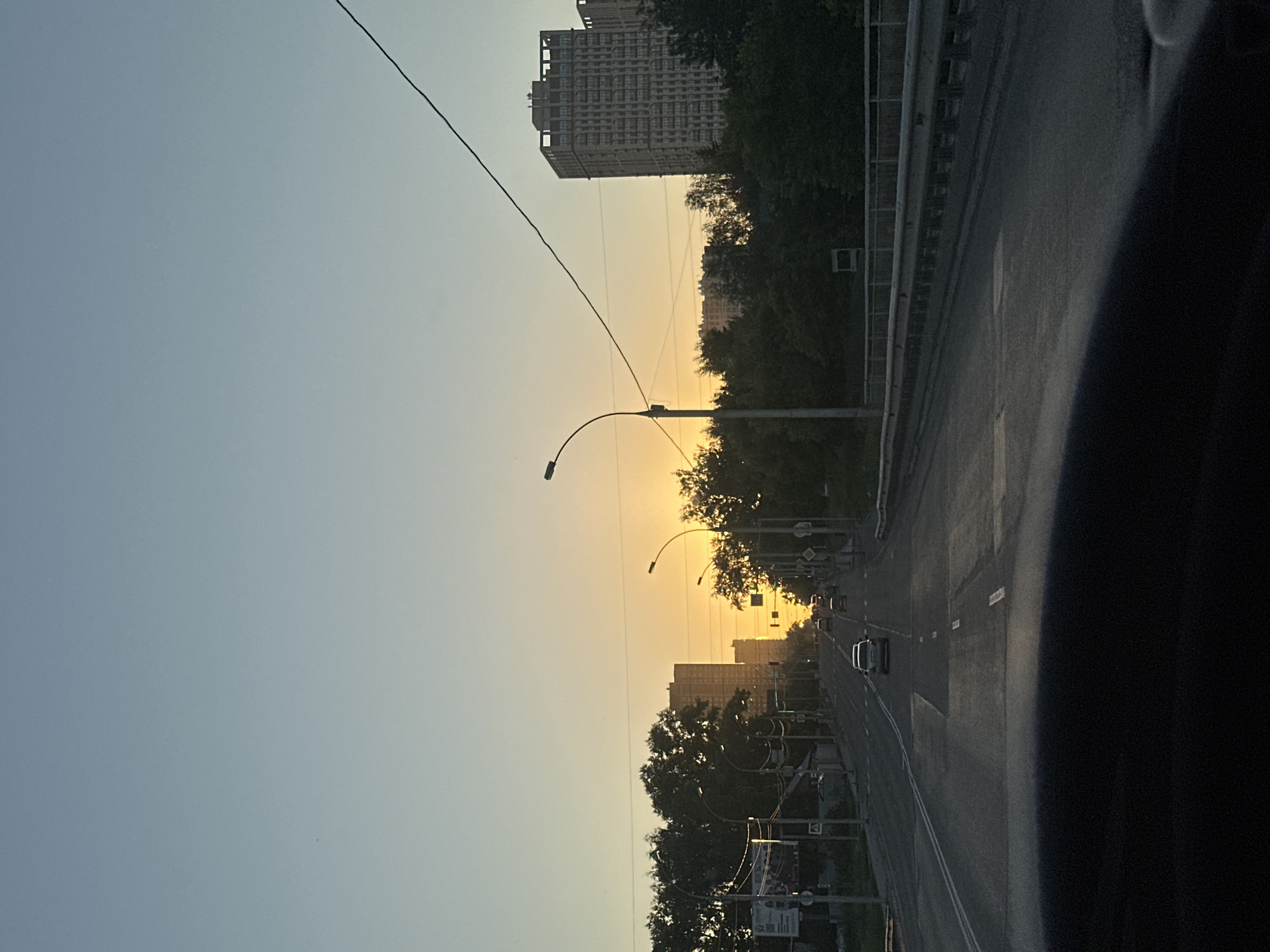 This is a vertical, eye-level, outdoor, full-shot image taken from inside a vehicle driving on a road towards the setting sun. The road appears to be a multi-lane highway with light traffic, including several cars and trucks. On either side of the road are trees and buildings, some of which appear to be apartment complexes. The sky is clear and a pale blue, transitioning to a bright orange and yellow hue near the horizon where the sun is setting. The overall impression is of a calm, urban scene at dusk. The presence of the multi-lane highway, the architecture of the buildings, and the specific lighting conditions suggest a location within the city of Novosibirsk, Russia, possibly along one of its major arteries leading out of the city center towards residential areas. The viewpoint is consistent with being on a bridge or elevated roadway, as indicated by the guardrail visible on the right side of the image.