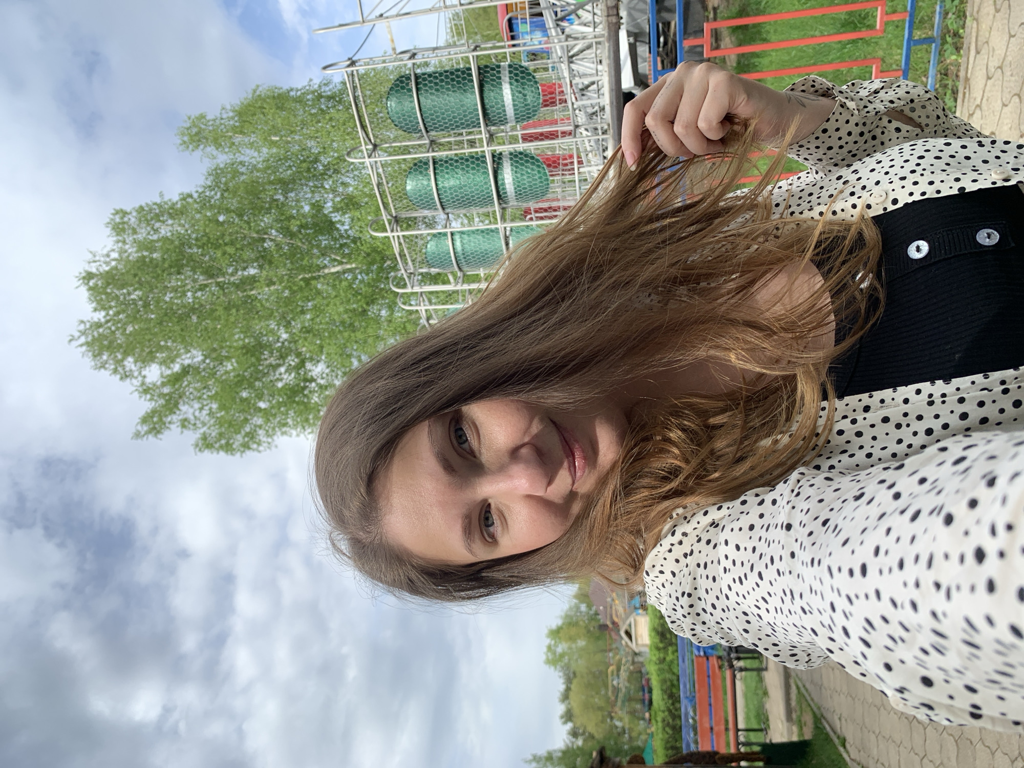 A woman poses for a selfie in Chaikovsky, Russia. The sky is cloudy. She is wearing a white and black polka dot blouse with a black vest. In the background is a tree, a metal structure with green canisters, and a fence.