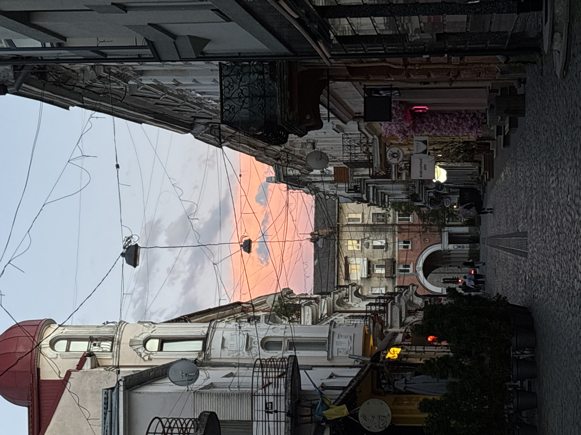 This image depicts a European-style street scene in Ternopil, Ukraine, likely during twilight. The perspective is looking down a cobblestone street lined with multi-story buildings on either side. The architecture features ornate detailing, balconies with wrought-iron railings, and arched windows. A prominent building on the left has a red domed roof and a curved bay window. On the right, similar buildings exhibit decorative elements and balconies.
The sky above is filled with dramatic clouds tinged with pink and orange, suggesting a sunset or sunrise. A complex network of overhead wires, likely for electricity and communication, crisscrosses the sky, with a few visible light fixtures hanging from them.
Several people are visible in the distance, walking along the street. Some are silhouetted against the fading light. The street itself is paved with cobblestones and has a visible drainage grate running down the center.
The ground level of the buildings appears to house shops and businesses, indicated by signage, awnings, and illuminated windows. One shop on the right is decorated with a large display of pink artificial flowers. Green potted plants are placed outside some establishments, adding a touch of greenery to the urban landscape. The overall impression is one of a historic city street in the evening.