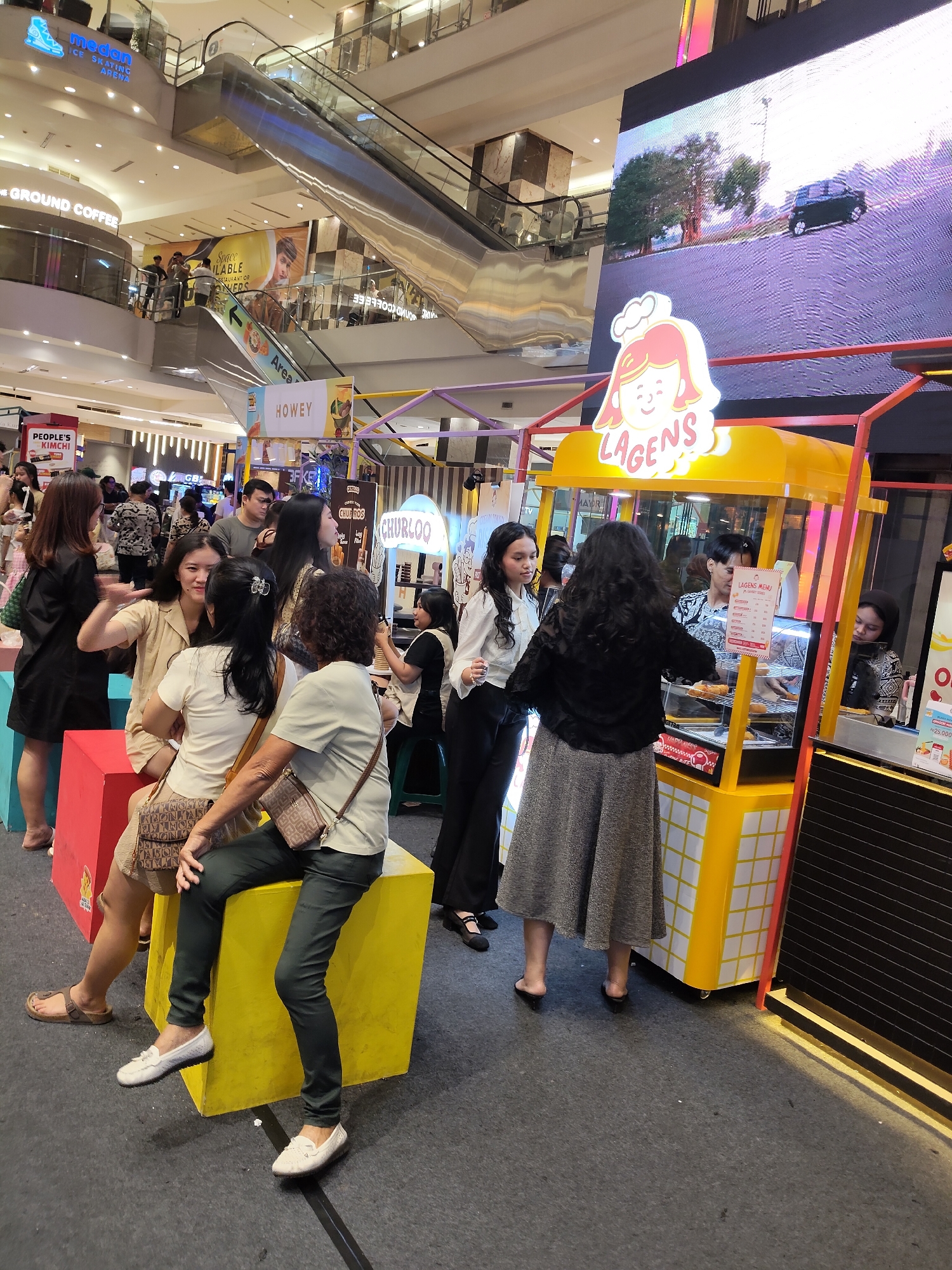 The image captures a bustling scene inside a shopping mall in the City of Medan, Indonesia. In the foreground, several women are interacting, with some seated on brightly colored blocks and others standing. One woman in a black dress is engaged in conversation with another woman in a beige outfit. Behind them, a bright yellow food stall with the sign "LAGENS" is visible, showcasing various food items in a display case. A menu board is also displayed at the stall, and a vendor appears to be serving customers. In the background, escalators are prominent, suggesting a multi-level shopping environment. A large digital screen displays a moving image of a car on a street. Other signs for businesses like "THE GROUND COFFEE" and "PEOPLE'S KIMCHI" are also present, indicating a commercial area. The overall atmosphere suggests a lively and active retail space.