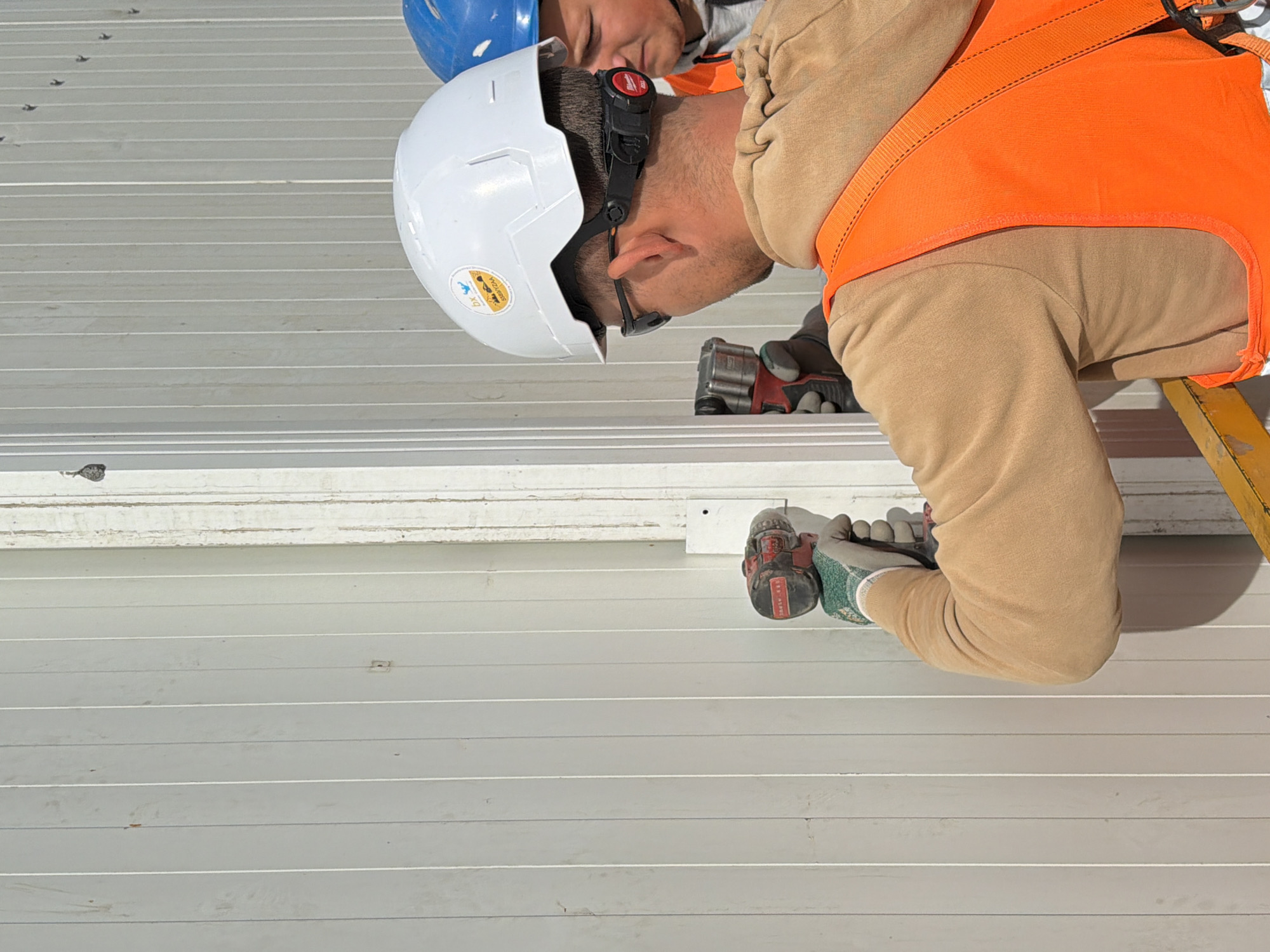 In Stróża, Poland, two individuals are working on an outdoor construction site, indicated by the bright sunlight and the construction materials. A man in the foreground, wearing a white hard hat, safety glasses, and an orange high-visibility vest over a tan hooded sweatshirt, is using a red and black cordless drill to attach a vertical white panel to a pre-existing structure. He is wearing green work gloves. Behind him, a second individual, also wearing a blue hard hat and an orange high-visibility vest, is looking on. The background consists of a white vertical siding structure, suggesting the construction of a building or fence. The overall scene depicts a typical construction activity.