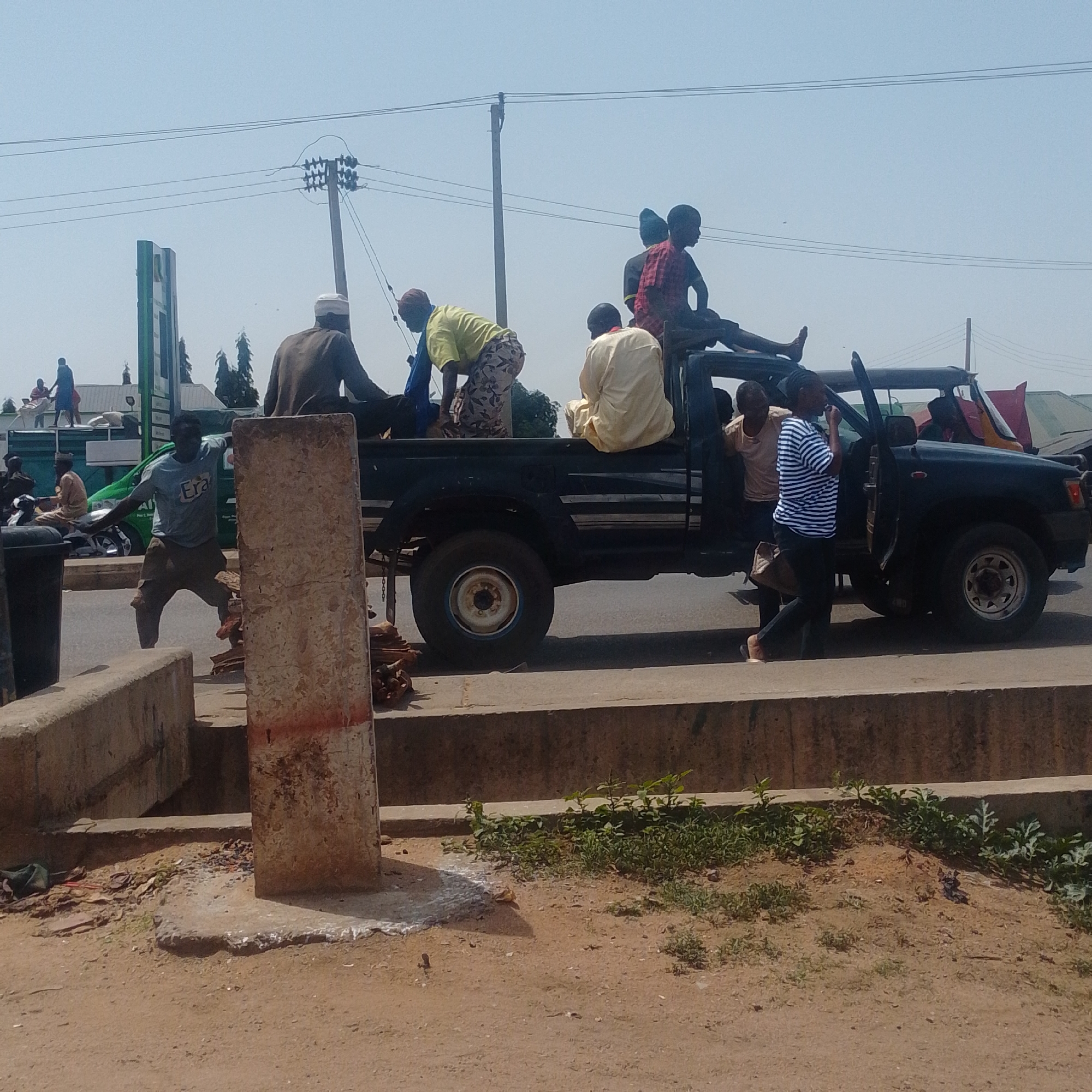 In Tumfure, Nigeria, a black pickup truck is parked on a paved road with several individuals riding in the truck bed and near the open driver's side door. The truck is filled with passengers, some sitting on the edge of the truck bed, others in the center. A few people are standing outside the truck. In the foreground, a concrete barrier runs along the edge of the road, with a concrete pillar or post in front of it. The environment appears to be an urban or semi-urban setting with overhead power lines and a clear sky. The people are dressed in casual clothing. The scene suggests transportation of people, possibly in a crowded manner.