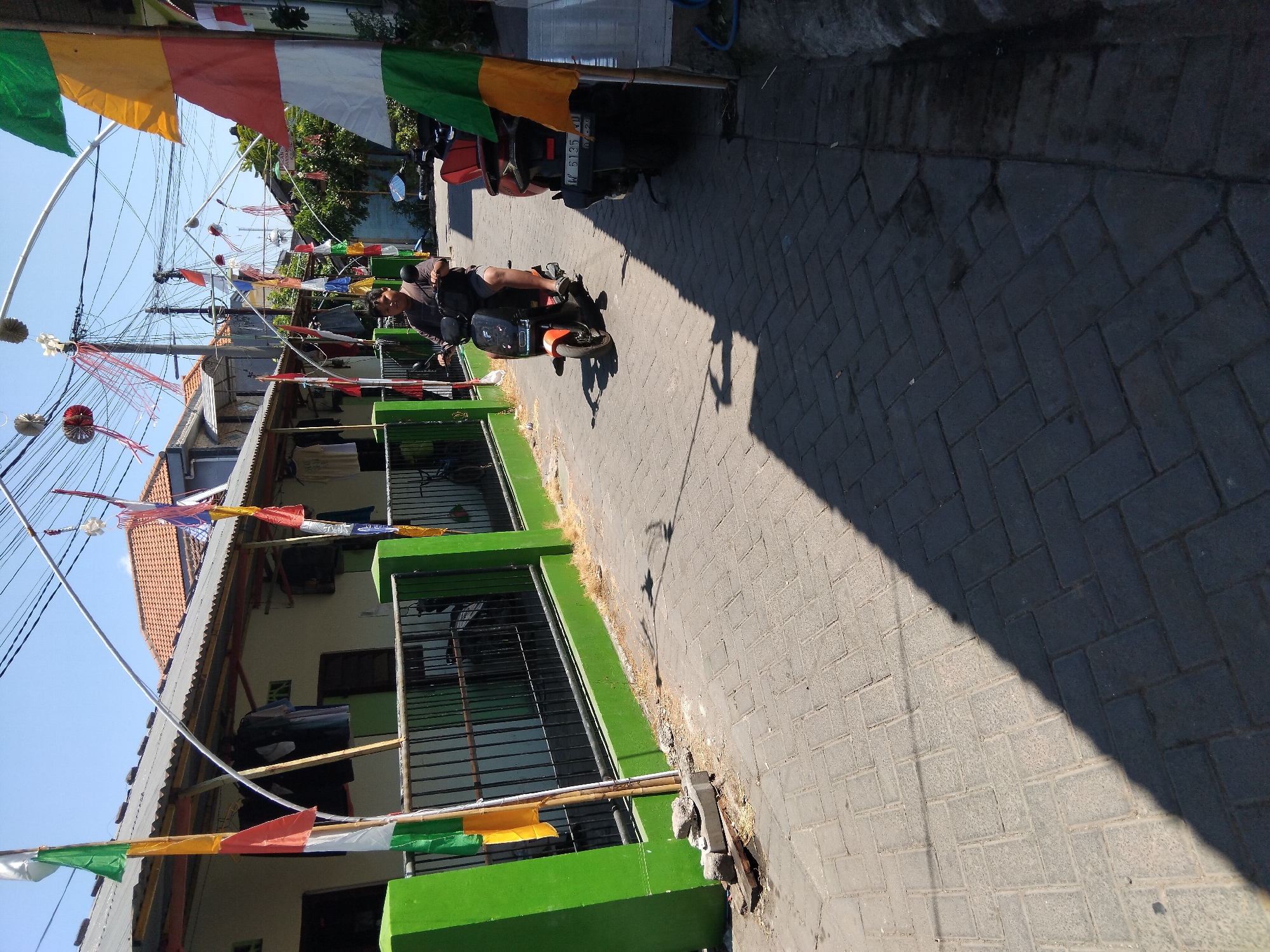 A man rides an orange electric scooter down a narrow, paved alley in Kepuh Kiriman, Indonesia. The alley is lined with houses, their walls painted a light color and their roofs made of corrugated metal. Colorful flags, red, white, and green, hang from poles along the alley, adding a festive atmosphere. Laundry is visible hanging outside one of the houses. The sun is bright, casting long shadows across the pavement. Another scooter is parked on the right side of the alley. The sky is a clear blue.
