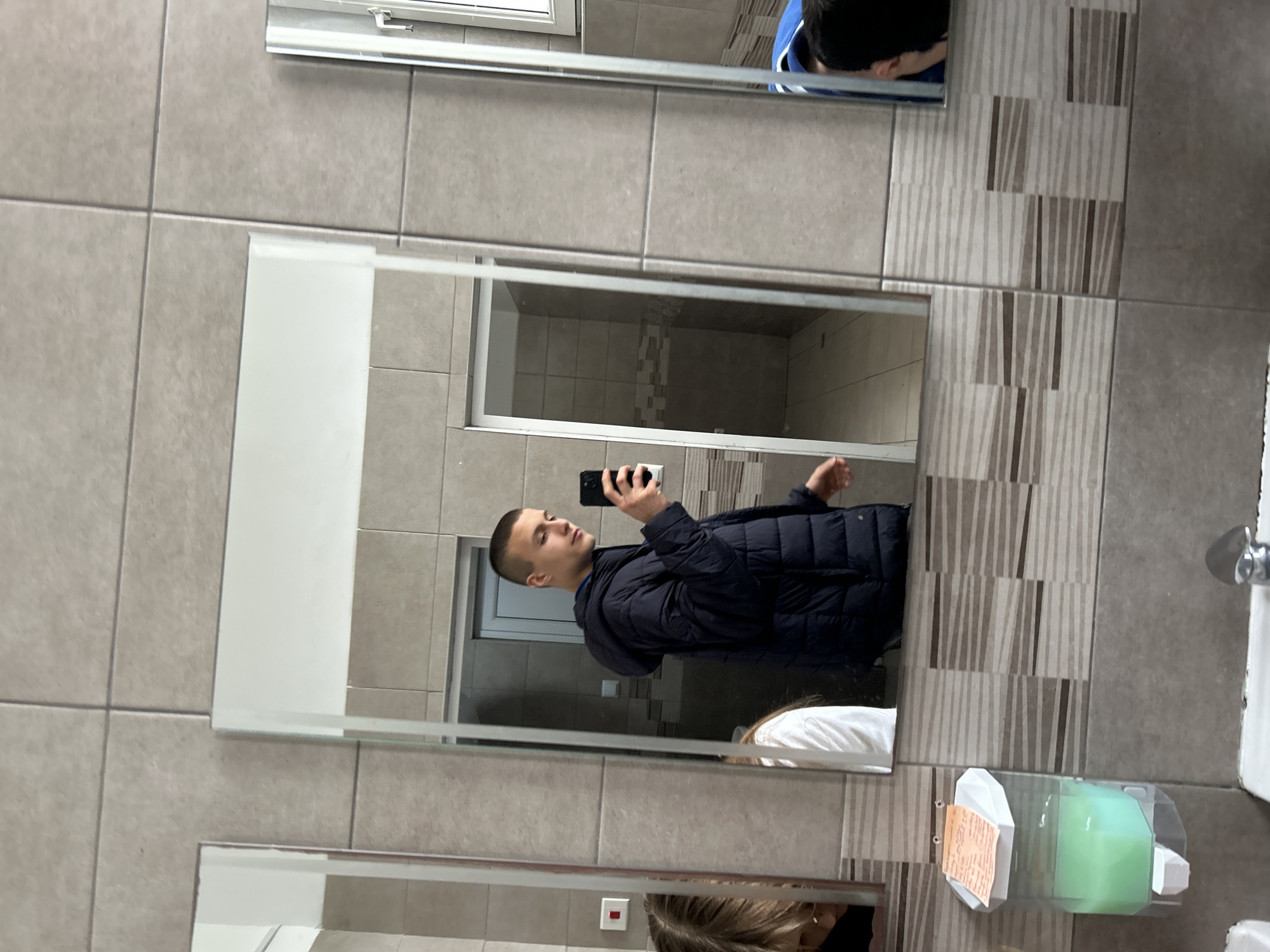 A young, light-skinned male with short hair is taking a selfie in a restroom mirror. He wears a dark blue puffer jacket over a blue shirt. The restroom has light gray square tiles on the walls and alternating vertical stripes of light gray and tan tiles. A soap dispenser and hand towel holder are visible. The restroom may be located in Burgas, Bulgaria but there is nothing in the image to verify that.