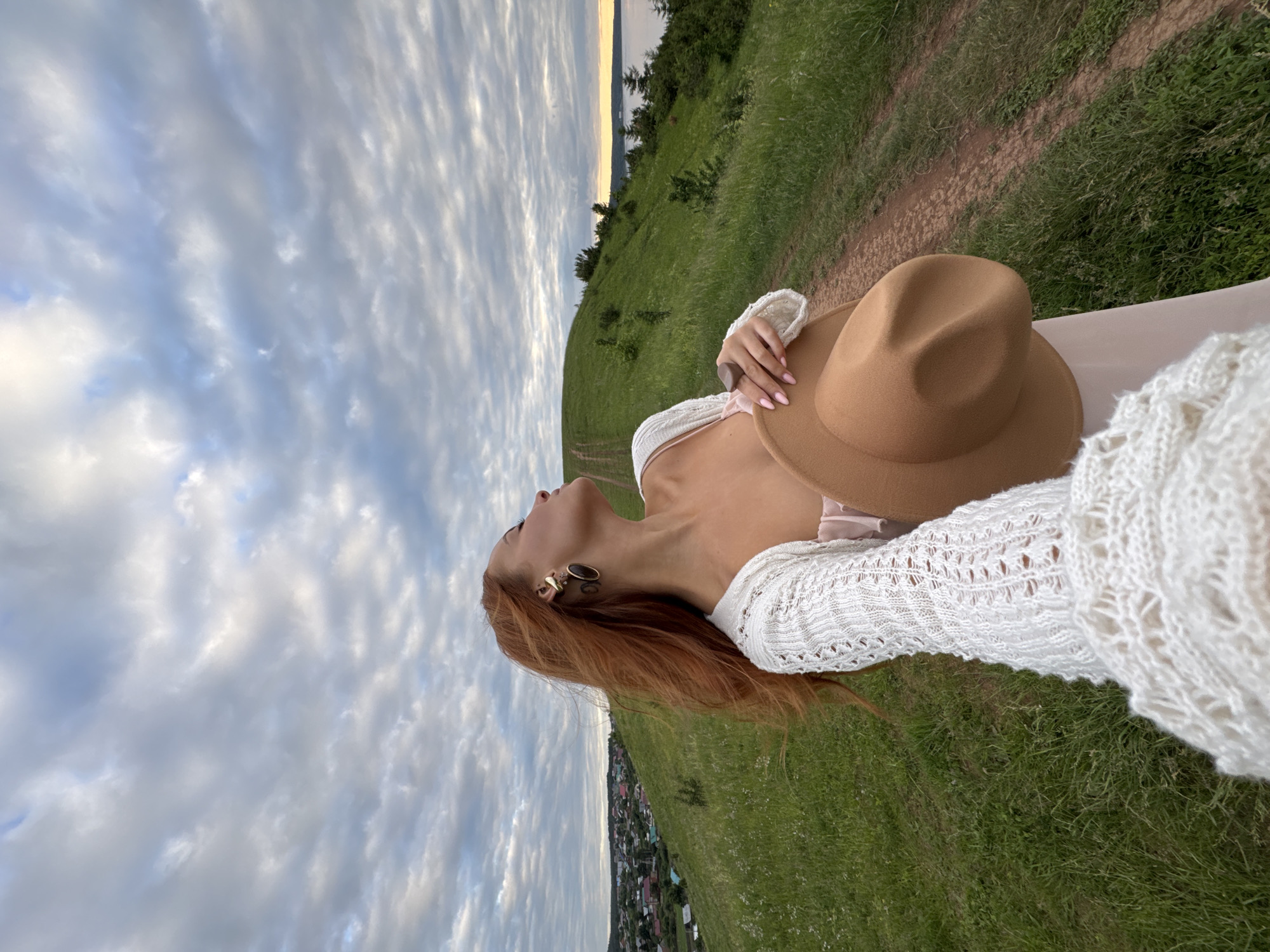 A woman with long, auburn hair is holding a light brown hat in her right hand, wearing a white knitted cardigan over a light pink dress, and large, dark-toned earrings. She is standing on a grassy hill with a dirt path winding up it. In the distance, a small village with scattered houses and a body of water can be seen. The sky is filled with cloudy, overcast skies. The image was taken in the city of Olkhovka, Russia.