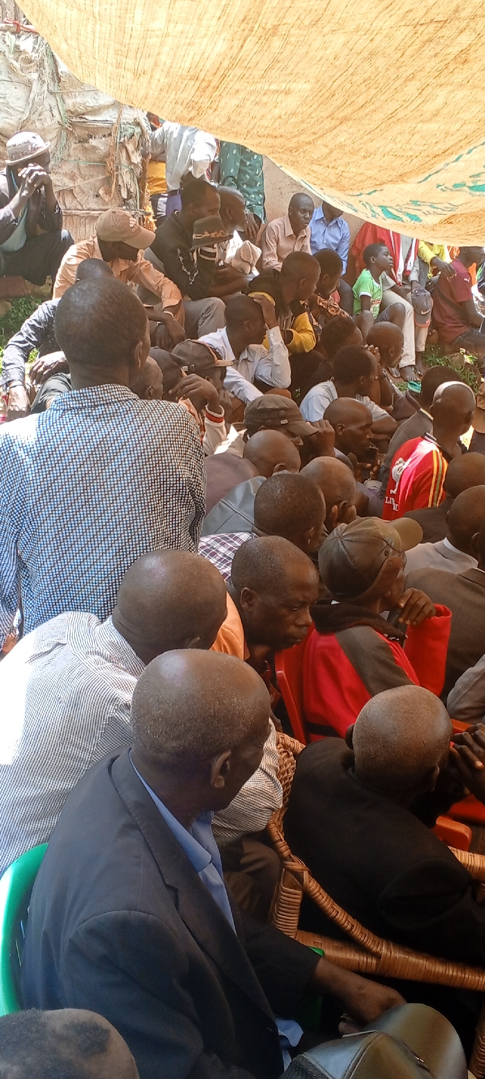 The image displays an outdoor community gathering in Kundu 1, Democratic Republic of the Congo. Approximately 30-40 individuals, primarily adult males of African descent, are densely assembled under a large, light-tan, woven fabric canopy providing shade. Most individuals are seated or squatting, facing generally forward towards an unseen point beyond the frame. Seating includes wicker chairs, green plastic chairs, and direct occupancy on the earth ground. Attendees wear diverse attire, including plaid shirts, solid-colored collared shirts, t-shirts, and some jackets; headwear like baseball caps and traditional-style caps is also observed. Sparse green vegetation is visible on the ground towards the right. The background shows indistinct structures, possibly rudimentary shelters, made of natural materials.
