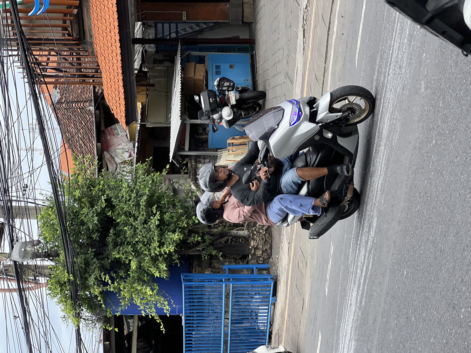 Two people are riding on a silver scooter on a street in Denpasar, Indonesia. The person in front is driving, wearing a black helmet, black long-sleeved shirt, and blue jeans shorts. They are holding the handlebars and have a ring on their left hand. The person behind is wearing a black helmet, a light pink long-sleeved shirt, and light blue pants. They are seated behind the driver. The scooter has a visible license plate that reads "DK 2339 ABY". The scooter has a black storage box on the back.
In the background, there is a bright blue metal fence to the left, and a large green tree behind it. Further in the background, there are buildings with orange tiled roofs, and numerous overhead electrical wires and poles. A second scooter, black with a storage box, is parked on the side of the road near a blue structure with boxes stacked on top. The street surface is asphalt, with white lines indicating lanes. The sky is overcast. The overall environment appears to be a busy urban street.
