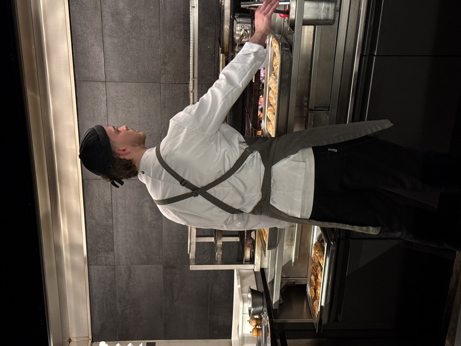 A chef in a white shirt and gray apron, wearing a black cap, stands in front of a large oven. Trays of baked goods are visible inside the oven. The chef's right arm is raised, appearing to adjust something within the oven. The kitchen setting includes gray tiled walls, stainless steel equipment, and black accents.