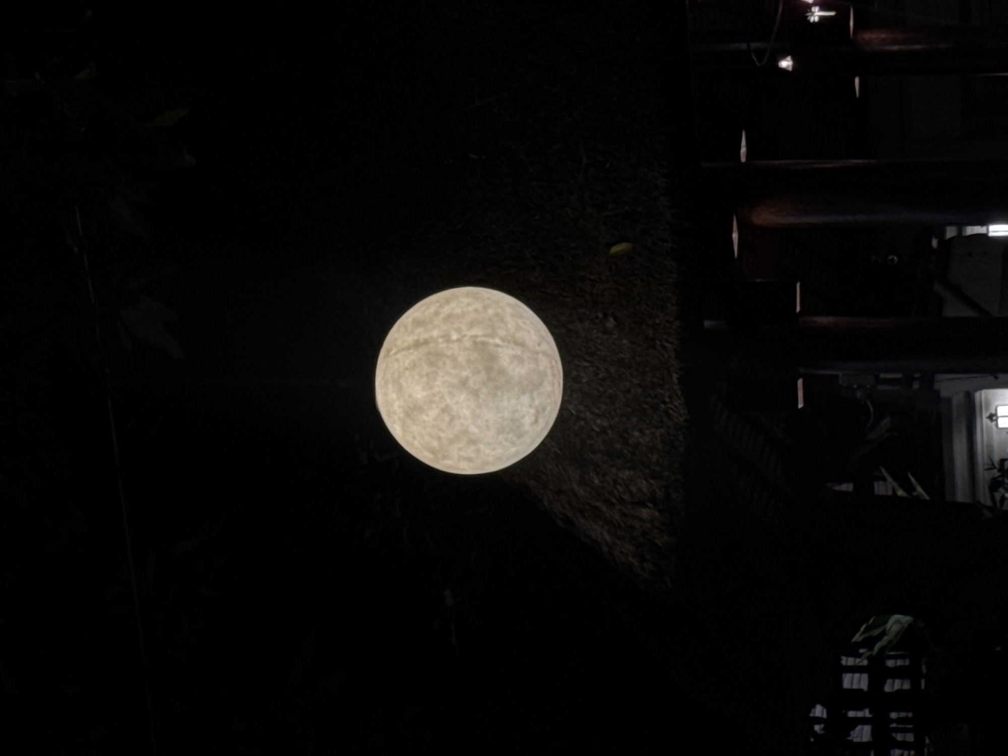 The image depicts a dimly lit outdoor scene, possibly at night, with a focus on a large, spherical light fixture hanging from above. The light source is illuminated with a warm, textured glow, resembling a full moon. Below the hanging light, a thatched roof structure is visible, suggesting an outdoor dining area or pavilion. The surrounding environment is dark, with silhouettes of foliage and architectural elements hinting at a tropical setting. In the lower portion of the image, the outlines of support columns and possibly tables and chairs can be discerned, further indicating a restaurant or bar setting. The overall impression is one of a serene, atmospheric evening ambiance. The context provided specifies the location as Kuta, Indonesia, which aligns with the visual cues of a tropical, possibly resort-style environment.