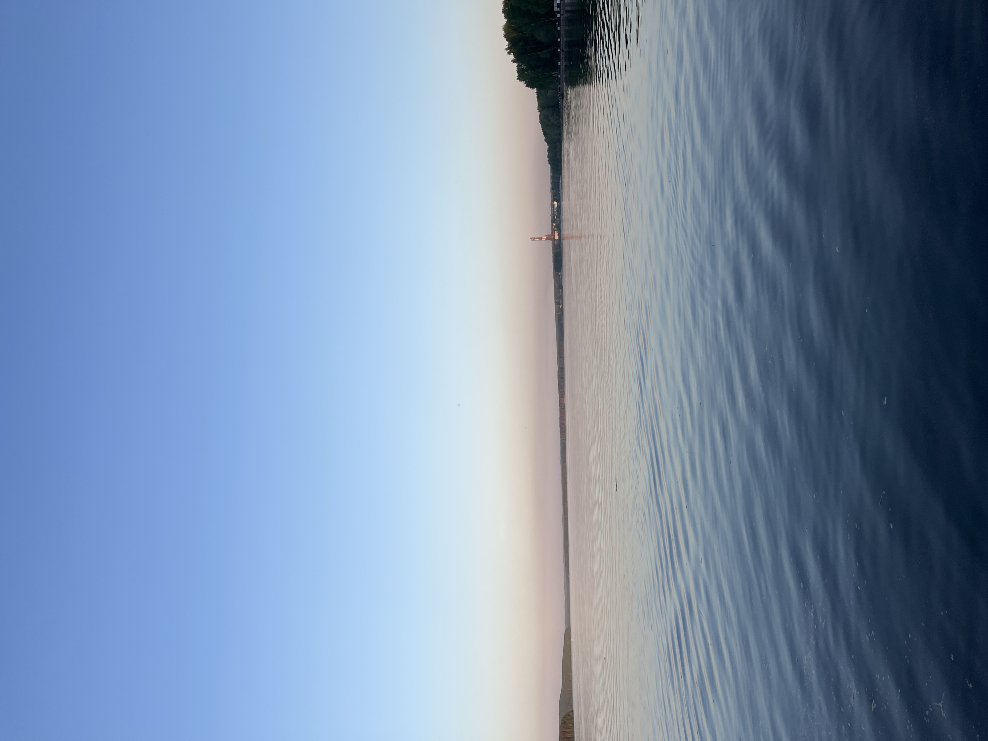 The image shows a body of water, likely the Kama River, in the city of Chaikovsky, Russia. On the far shore, a line of trees is visible, and a tall tower is visible. The sky is a gradient of blue, indicating either sunrise or sunset. The water is rippled, suggesting a light breeze.