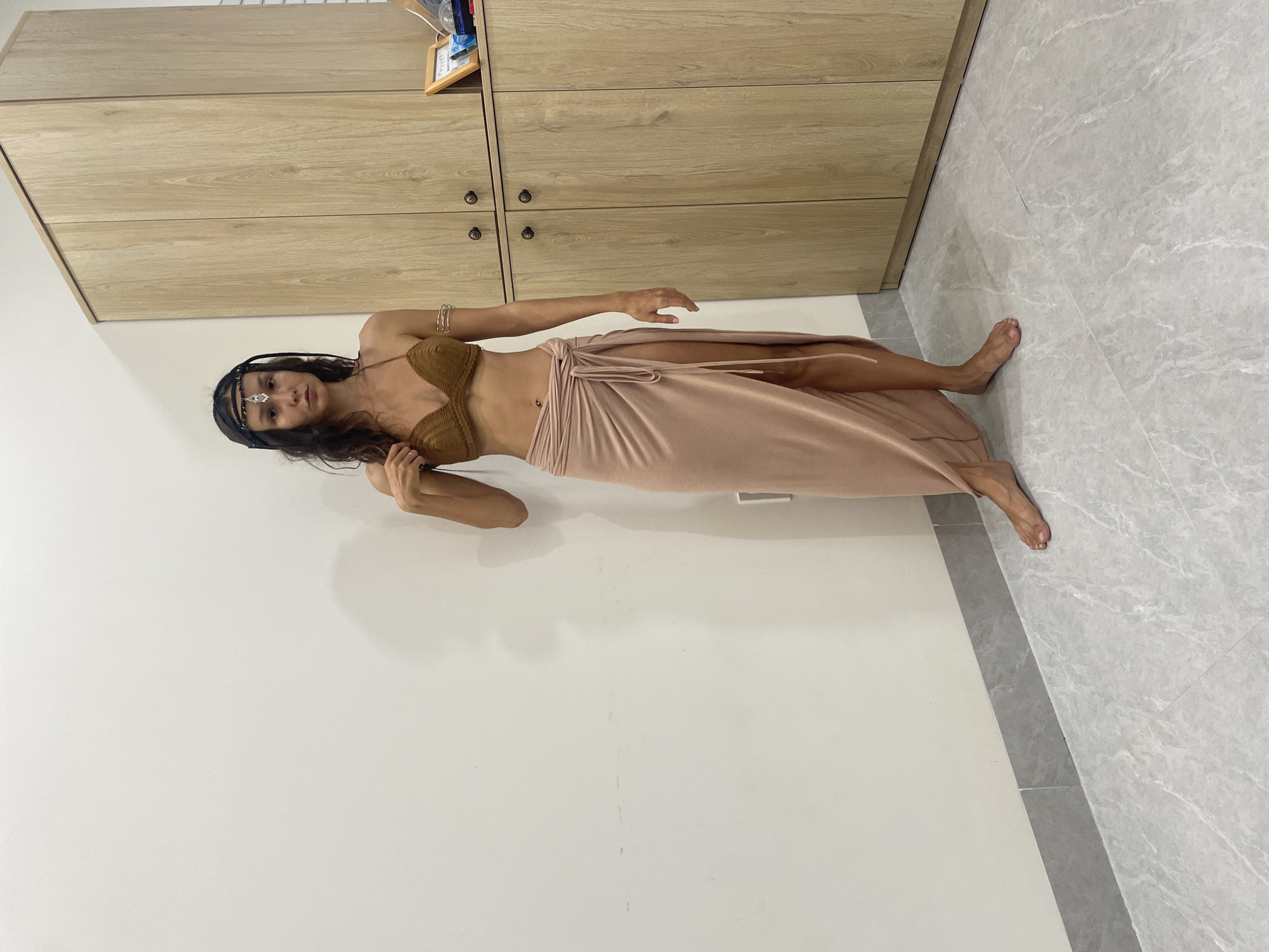 A woman stands in front of a plain white wall and a wooden cabinet in a room, possibly in Mũi Né, Vietnam. She has dark, wavy hair and is wearing a brown crocheted bikini top, a pale pink wrap skirt with a high slit, and a decorative headpiece. Her stance is relaxed, with one hand touching her chest and the other resting by her side. The floor is tiled with a gray marble pattern. The wooden cabinet appears to be a wardrobe or storage unit with multiple doors and handles. A framed picture and some small objects are visible on top of the cabinet. The lighting suggests an indoor environment with artificial light.