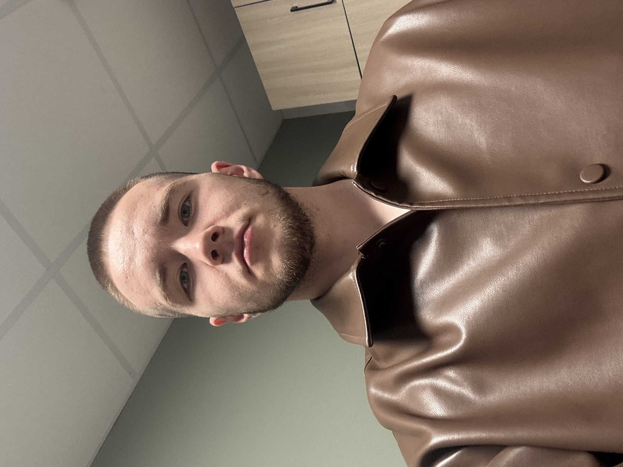 The image shows a young man taking a selfie indoors. He has light skin, light-colored eyes, very short light brown hair (buzzed style), and a short, neatly trimmed beard. He is looking directly into the camera with a neutral expression.
He is wearing a brown jacket, which appears to be made of leather or a similar material, with a slight sheen. The jacket has a collar and a visible round button on the right side of his chest.
The background suggests an indoor setting, possibly an office or public building. The wall behind him is a plain, light grey-green color. Above him, a ceiling with visible square tiles and seams can be seen, which is characteristic of institutional or commercial architecture. In the upper left background, part of a wooden or laminated cabinet with a dark horizontal handle is visible, adding to the indoor environment.
The photograph is oriented sideways, giving the impression that the man is lying down or the camera was held at a 90-degree angle. The lighting is artificial, casting some soft shadows on his face and the folds of his jacket. There is no visible text in the image. The provided context indicates the location is Kazan, Russia.