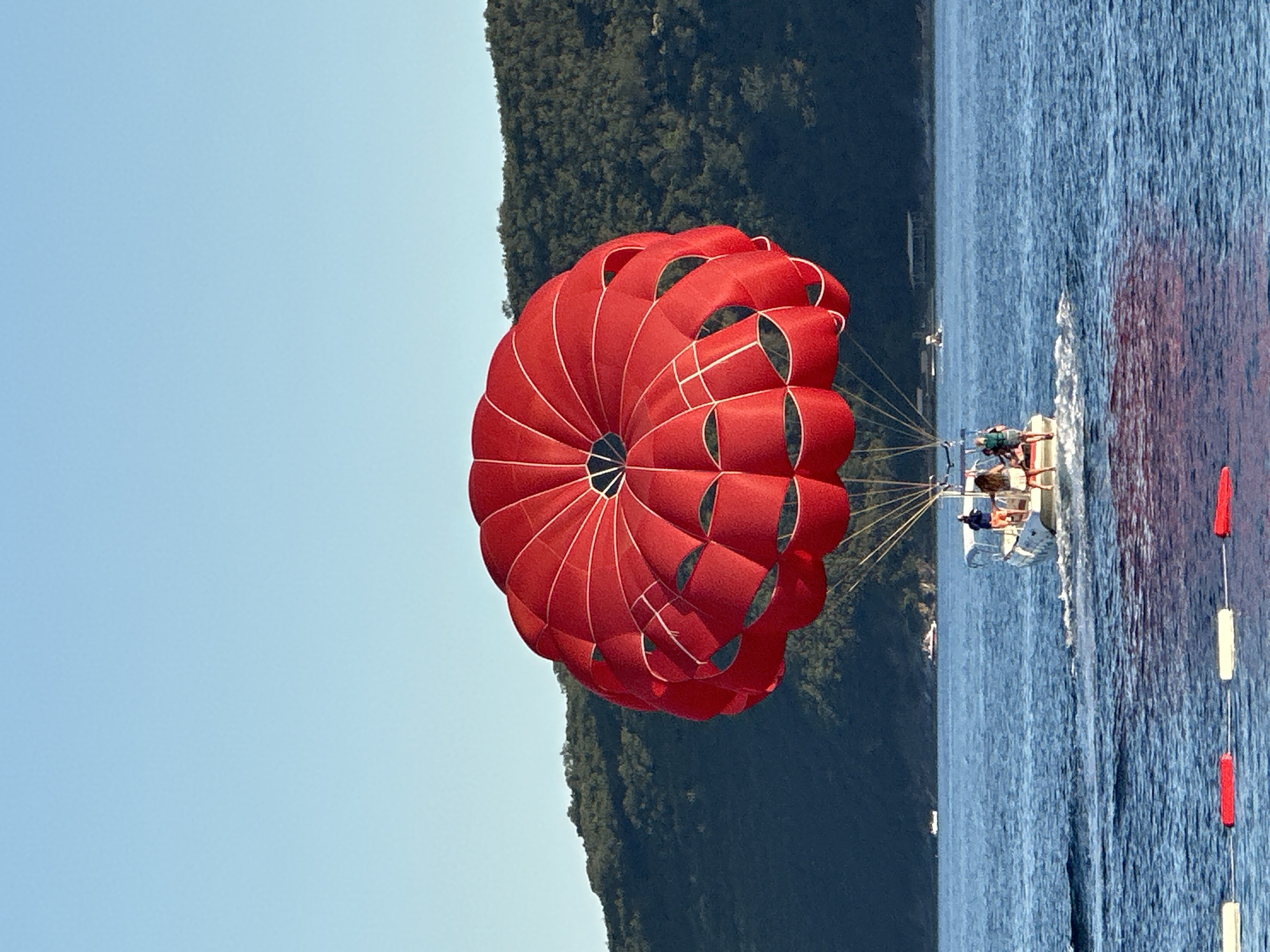 A bright red parasail is tautly inflated, billowing above a white motorboat on a deep blue sea. The boat, moving at speed and creating a white wake, carries four people. Two individuals stand at the stern, appearing to be holding ropes connecting them to the parasail. One person is seated behind them, and another stands near the front. In the background, a verdant, tree-covered hillside rises sharply from the coastline. The sky above is a clear, pale blue. The scene appears to be taking place in a bay or coastal area near Budva, Montenegro, given the hilly, Mediterranean landscape. Floating buoys, alternating red and white, are visible in the foreground, marking a boundary on the water.