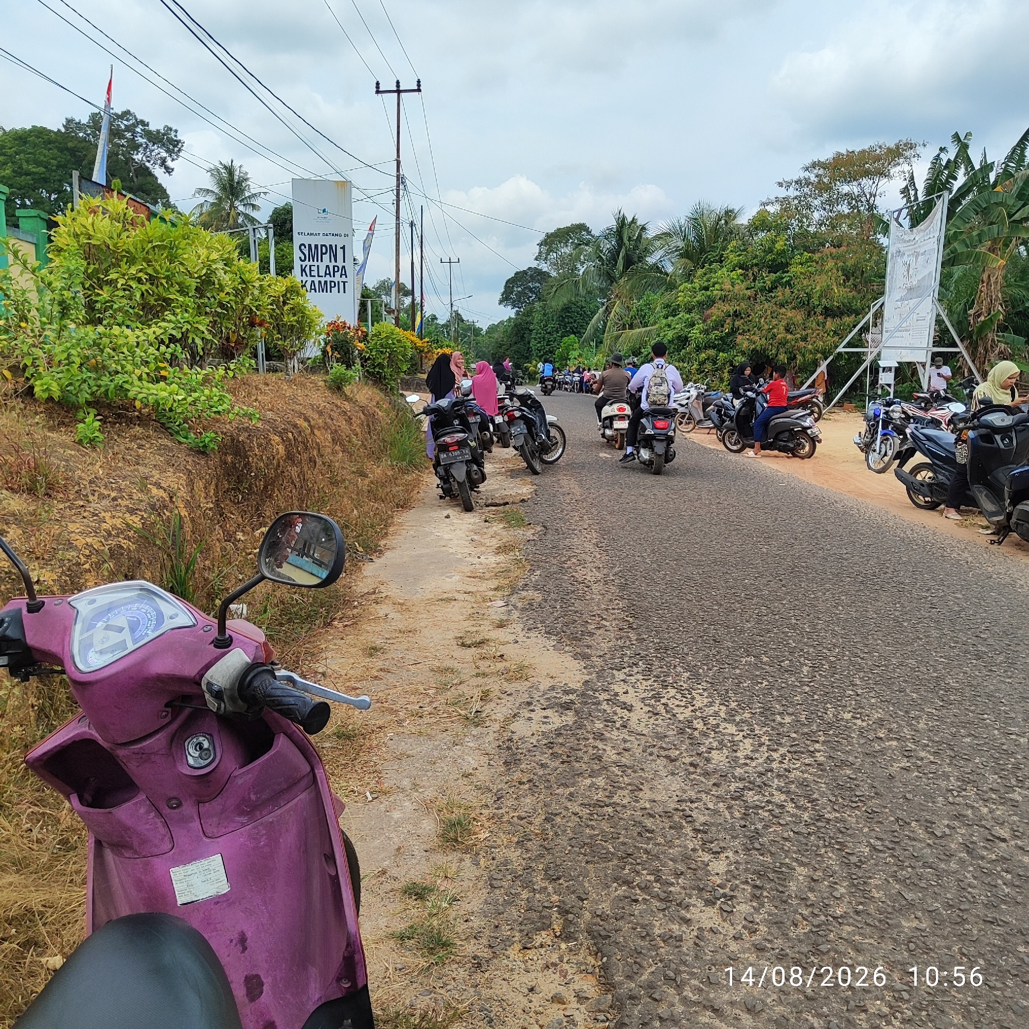 An outdoor, eye-level, medium shot captures a dirt road in front of a school. On the left, a purple scooter is parked on the side of the road, its front end facing the camera. To the right of the scooter, the road is lined with parked motorcycles and people. A white sign with "SMPN 1 KELAPA KAMPIT" is visible. The road is paved and appears to be well-traveled, with many motorcycles parked along its edges. The environment is rural, with lush green vegetation and trees in the background. The sky is overcast, with some clouds visible. The time and date stamp "14/08/2026 10:56" is present in the bottom right corner.