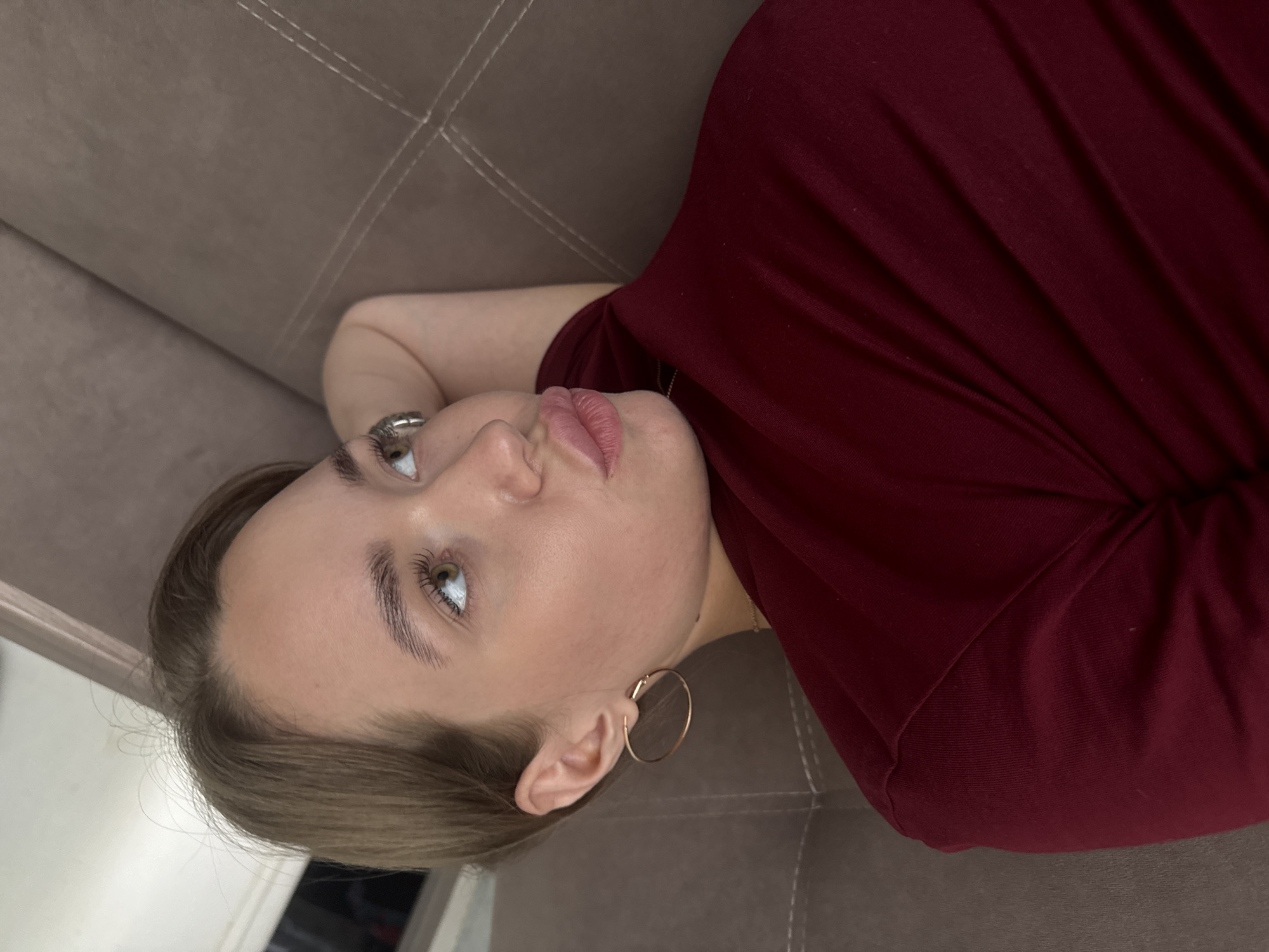 A young woman with blonde hair and hazel eyes is lying on a brown sofa. She is wearing a maroon-colored top and gold hoop earrings. Her mouth is slightly pursed, and she is looking away from the camera. The sofa appears to be in a residential setting, possibly a living room. The image does not provide any specific details about the location within the city of Chaikovsky, Russia, or any other geographical indicators.