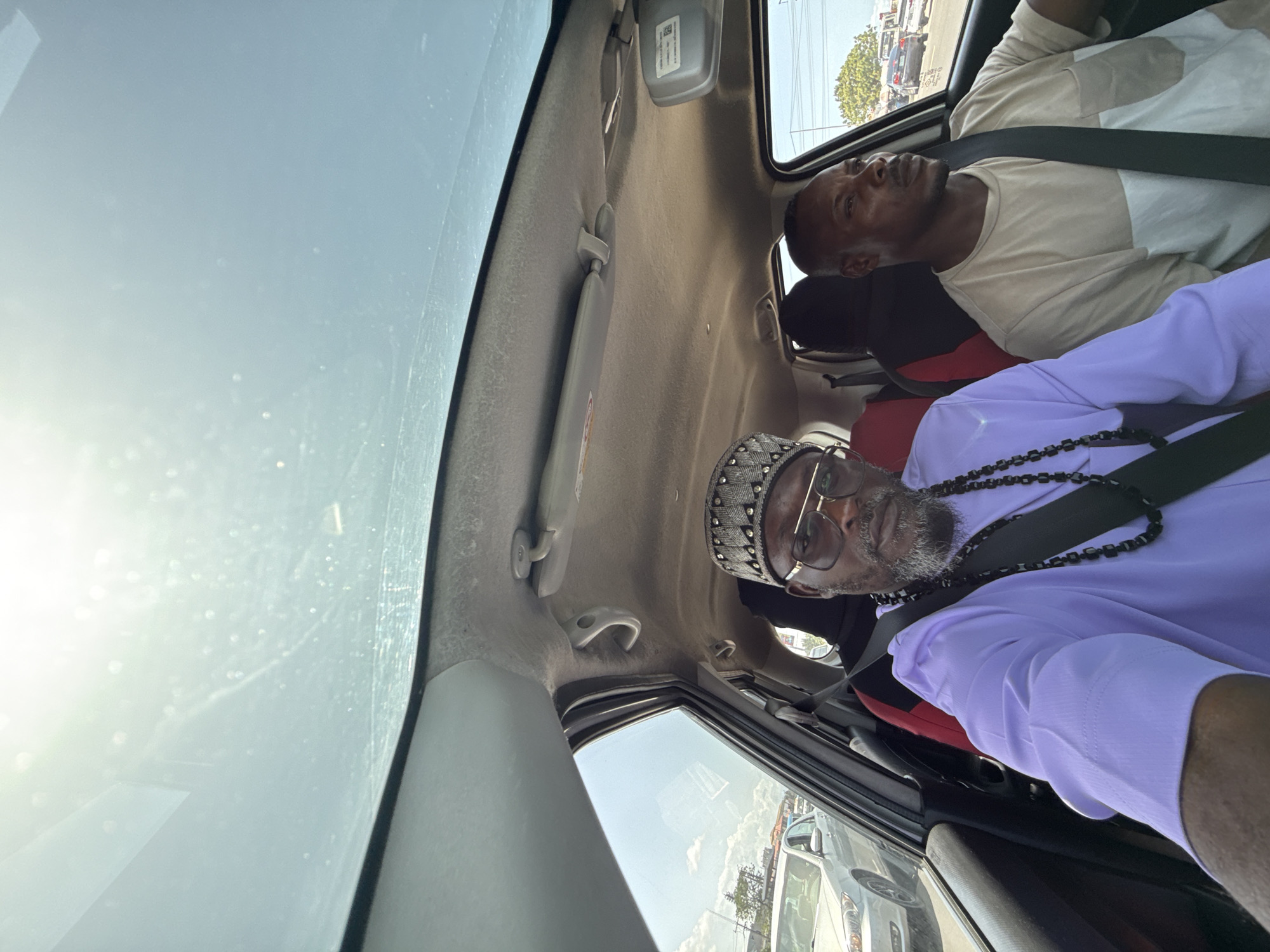The image shows two men in a car, both wearing seatbelts. One man in the front passenger seat wears a tan and white shirt and stares towards the front. The second man in the back passenger seat wears a lilac shirt, a beaded necklace, and eyeglasses. He also has a white cap on his head and a beard. The car has a sunroof. Outside the car, cars and buildings are visible, suggesting an urban setting in Ojomu, Nigeria.